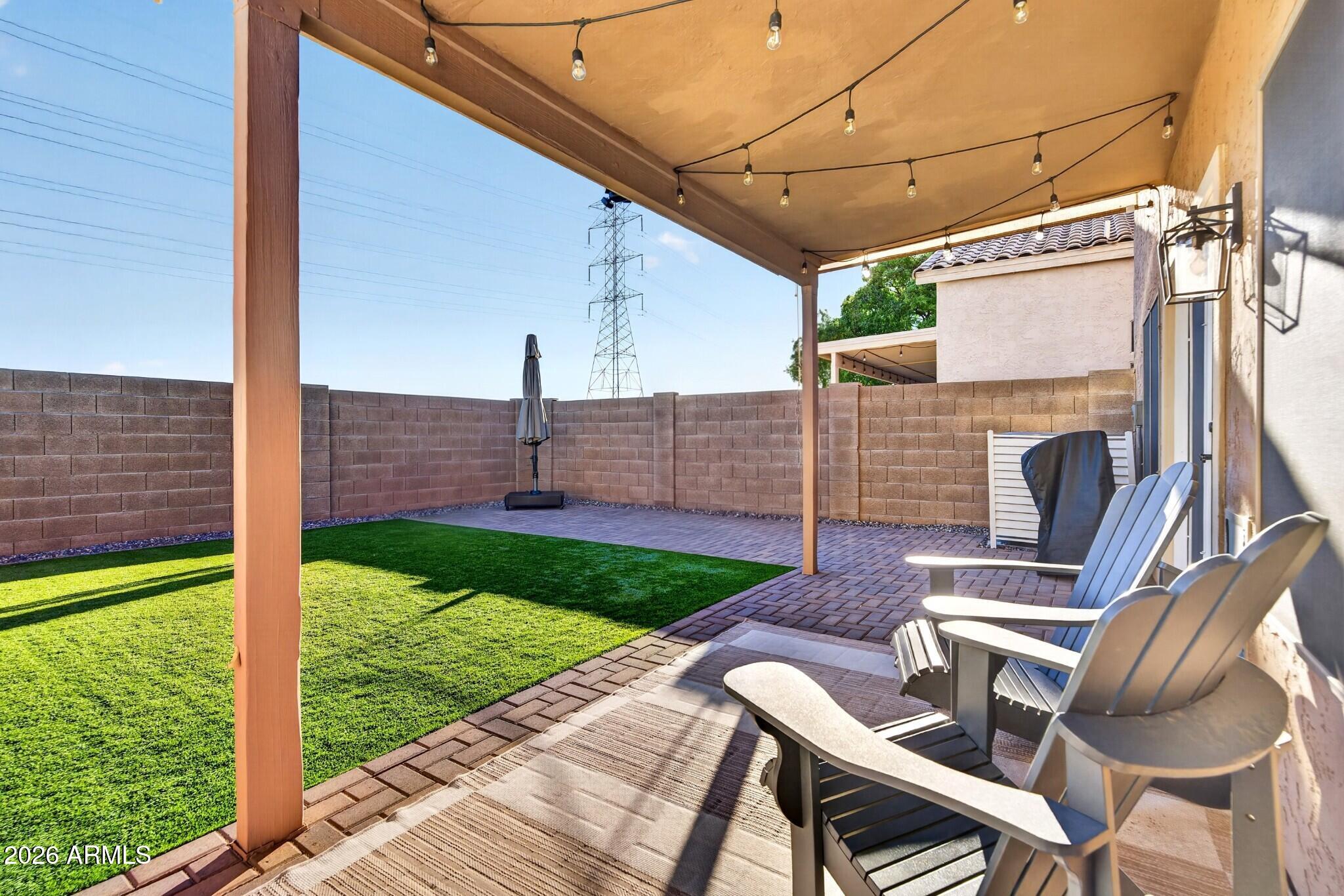 Covered backyard patio with string lights, Adirondack chairs, brick pavers, and a green lawn.