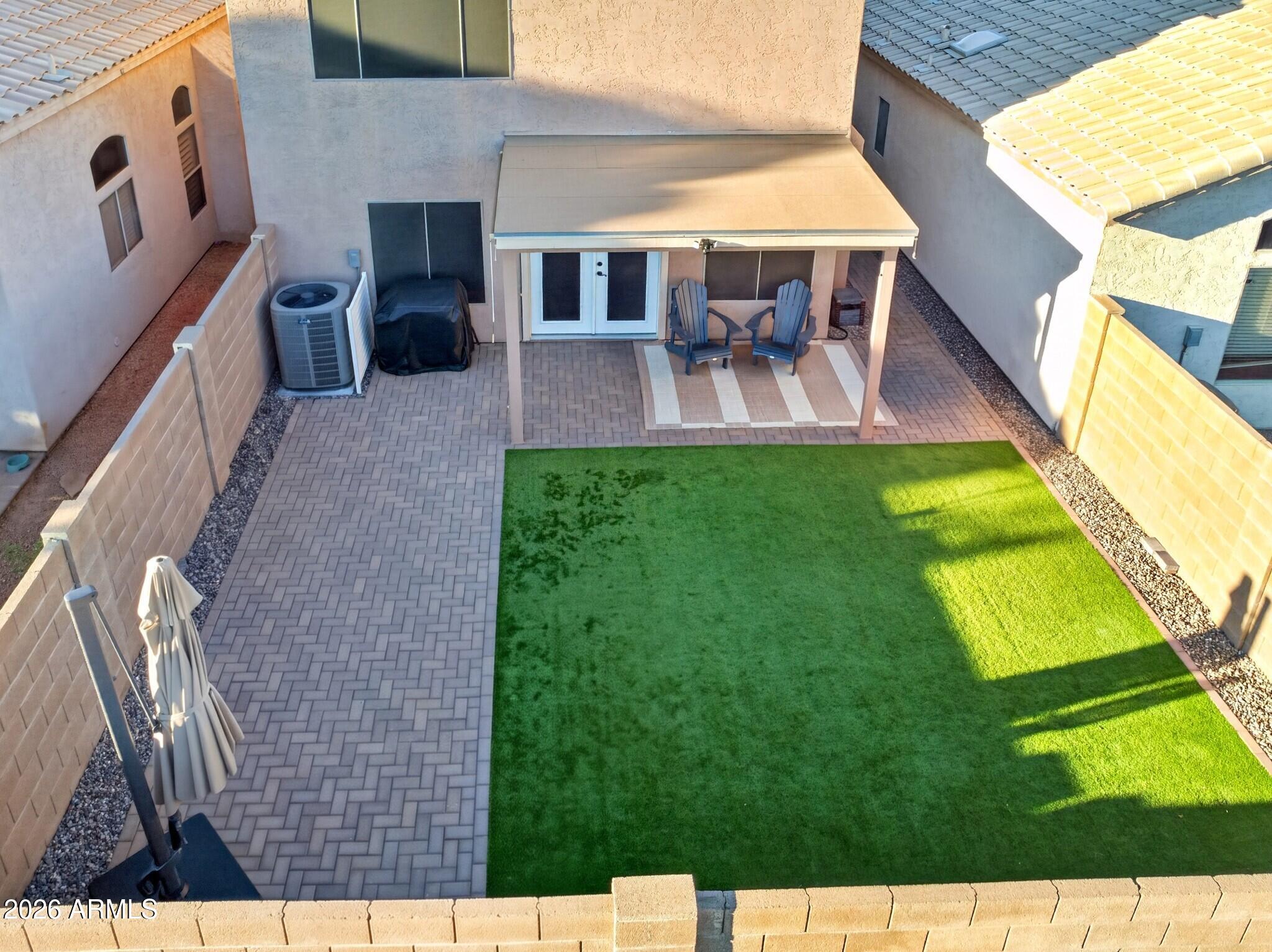 Walled backyard with artificial turf, paver patio, and covered seating area with two chairs.