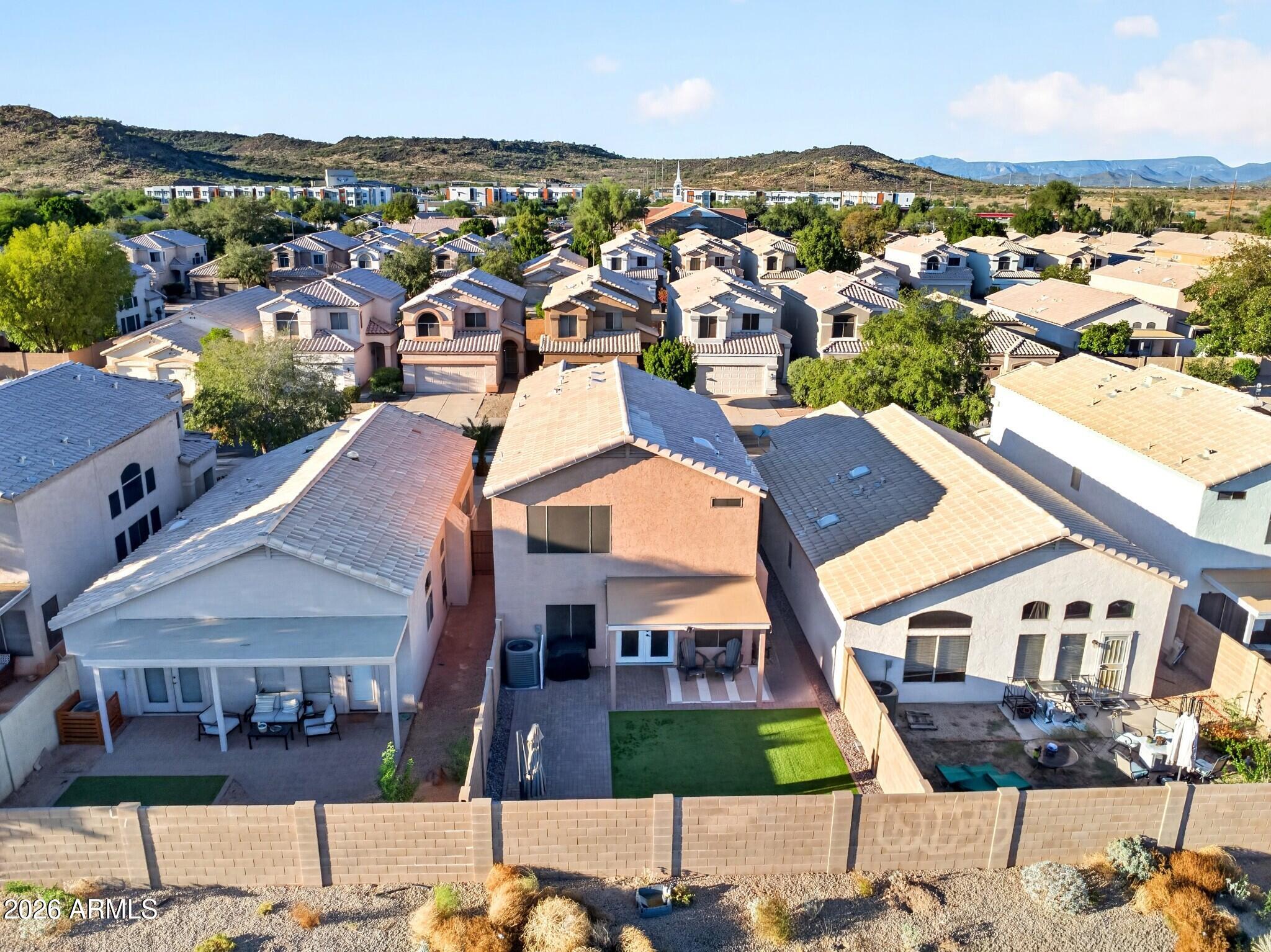 Aerial view of fenced backyard with turf, covered patio, neighboring homes, and distant mountains.