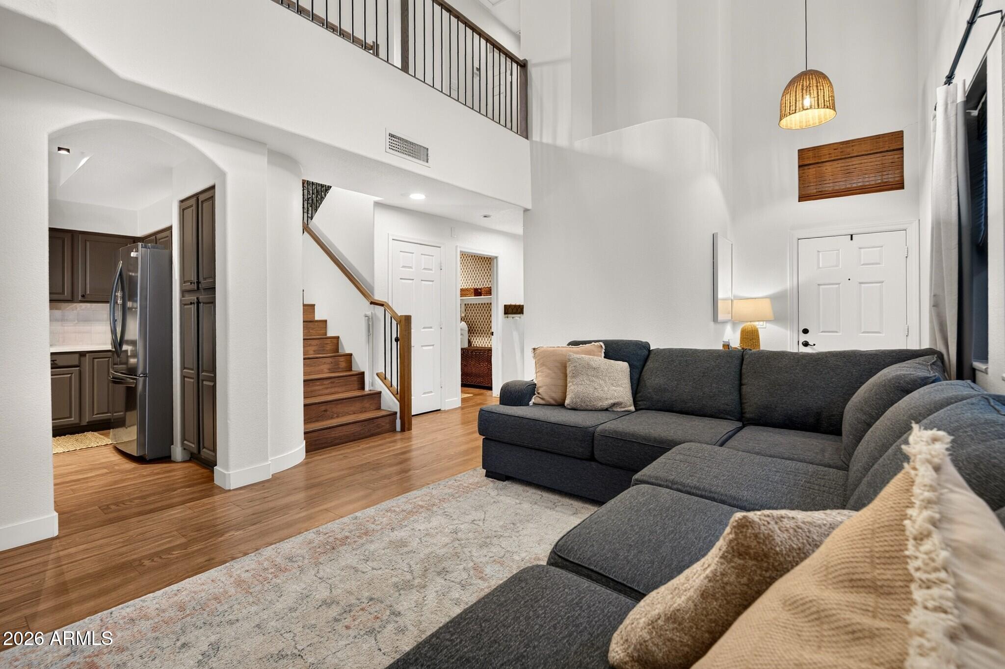 Double-height living room with a dark sectional, wood staircase, and kitchen visible through an archway.