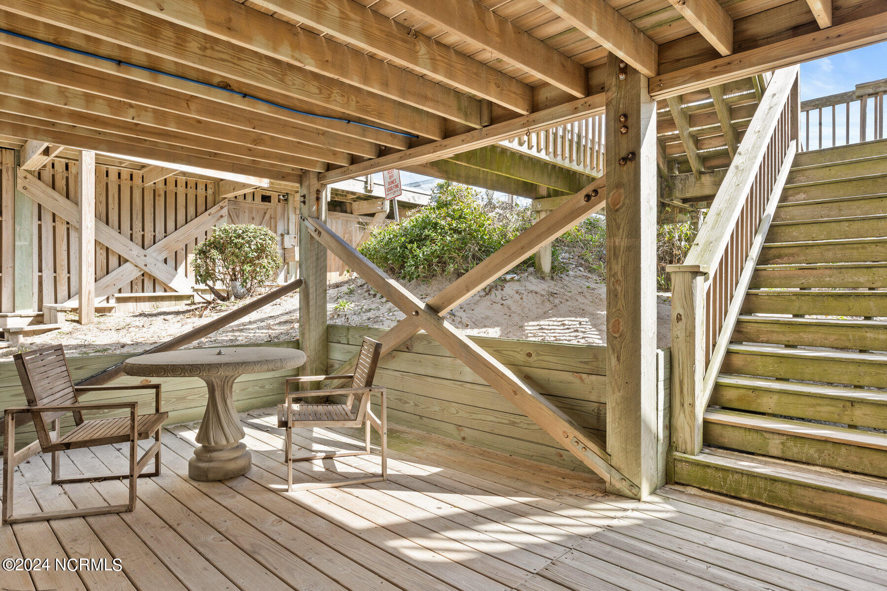 Covered ground-level deck with wooden chairs, a round table, and exterior stairs.