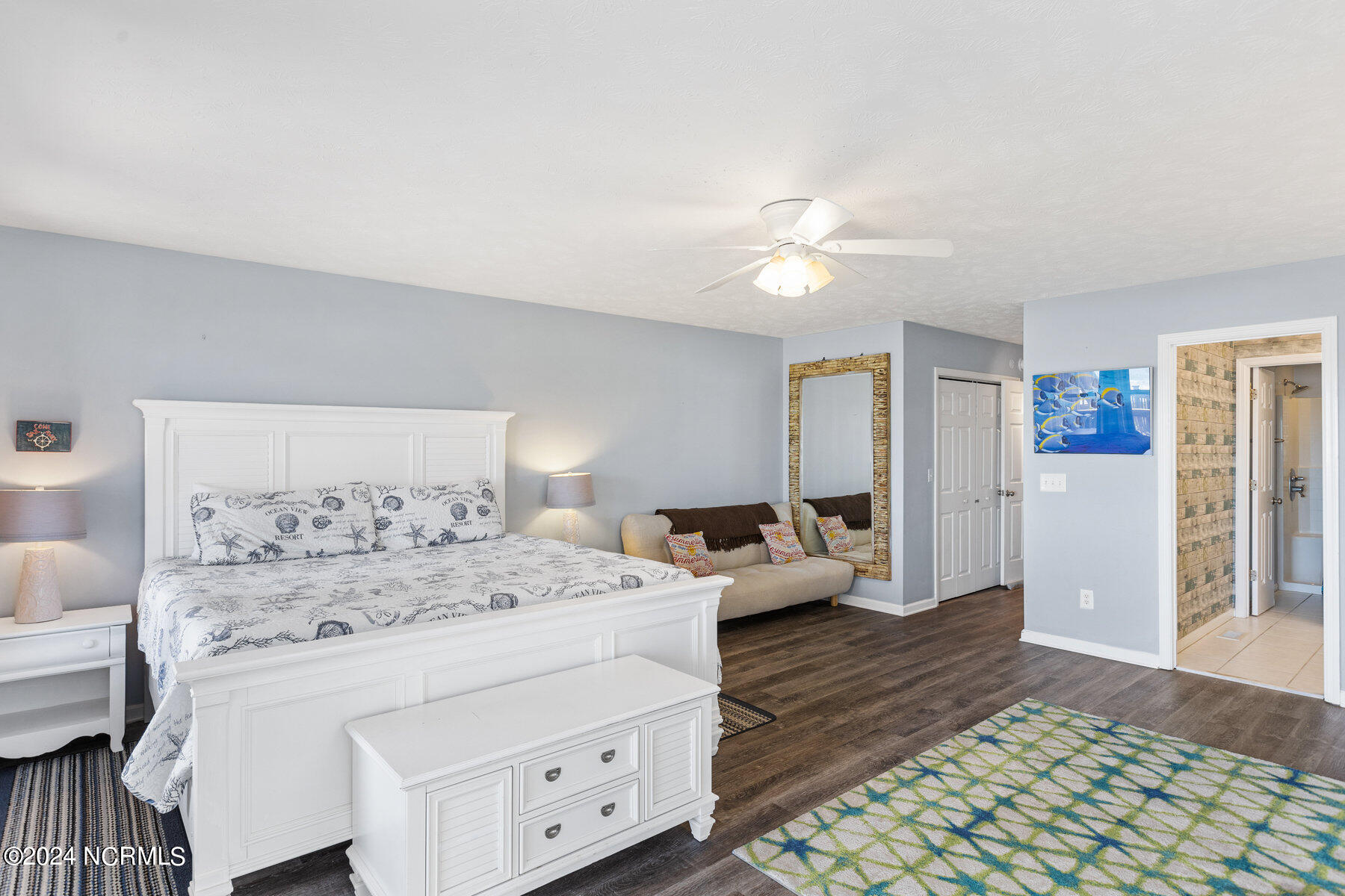 Furnished bedroom with a large white bed, loveseat, ceiling fan, and open doorway to a tiled bathroom.