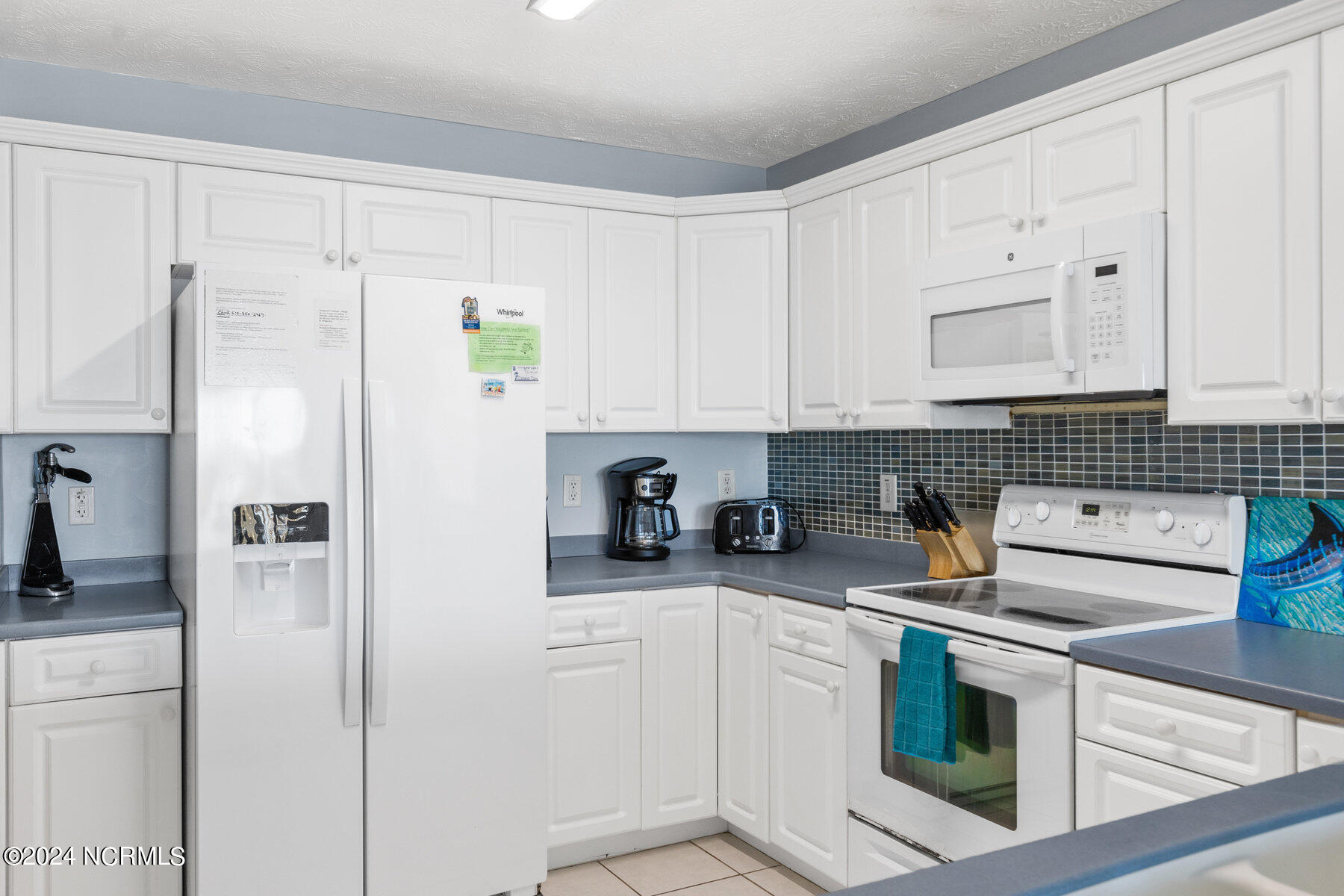 Kitchen with white cabinets, blue-gray counters, mosaic tile backsplash, and white appliances.