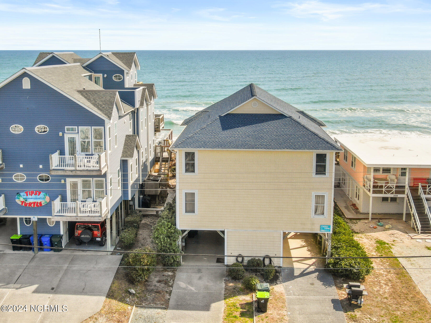 Pale yellow raised beach house beside the ocean, framed by blue and coral neighboring homes.