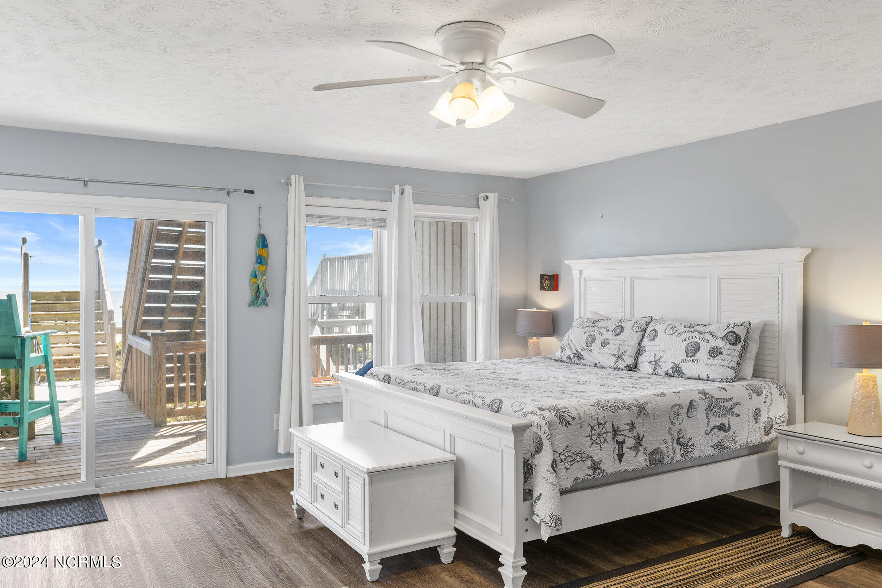 Furnished bedroom with coastal-print bedding and sliding doors opening onto a sunny deck.