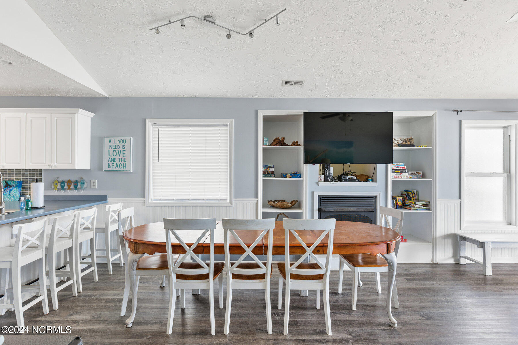 Dining area with a large wooden table, white chairs, and built-in shelves around a fireplace and television.
