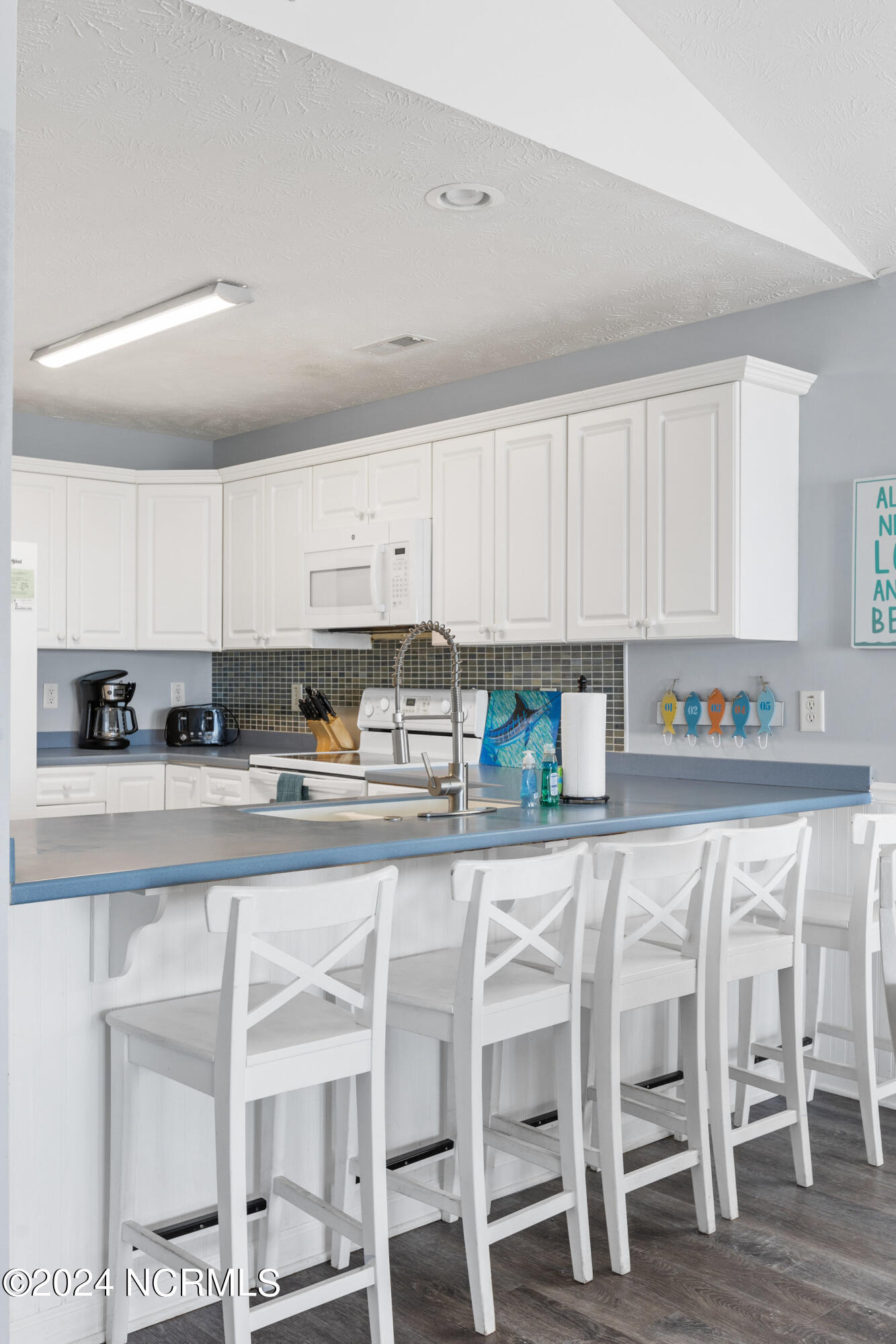 Kitchen with white cabinets, a blue-gray peninsula, white bar stools, and dark mosaic backsplash.