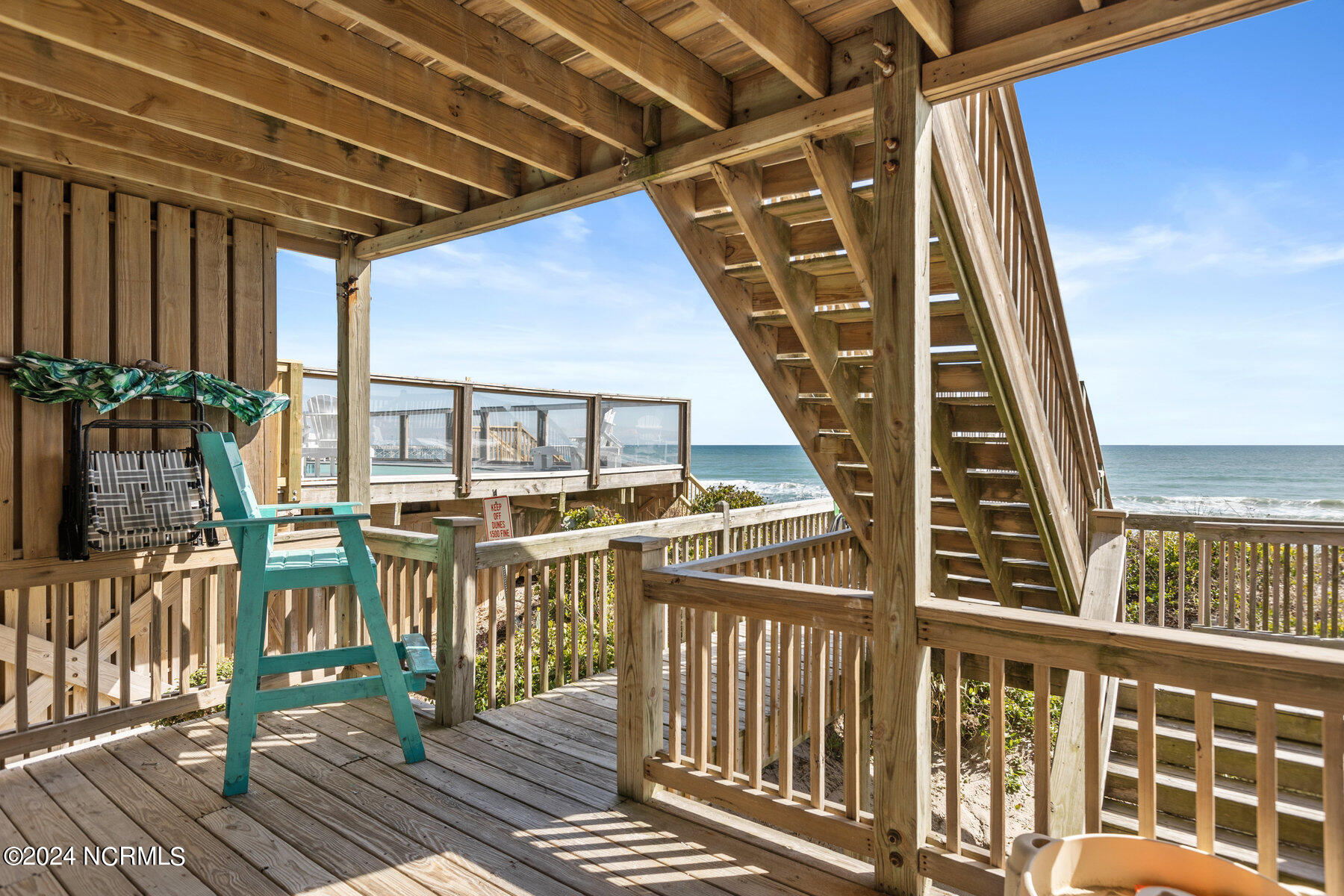Covered wooden deck and staircase overlooking the ocean, with a turquoise Adirondack chair.