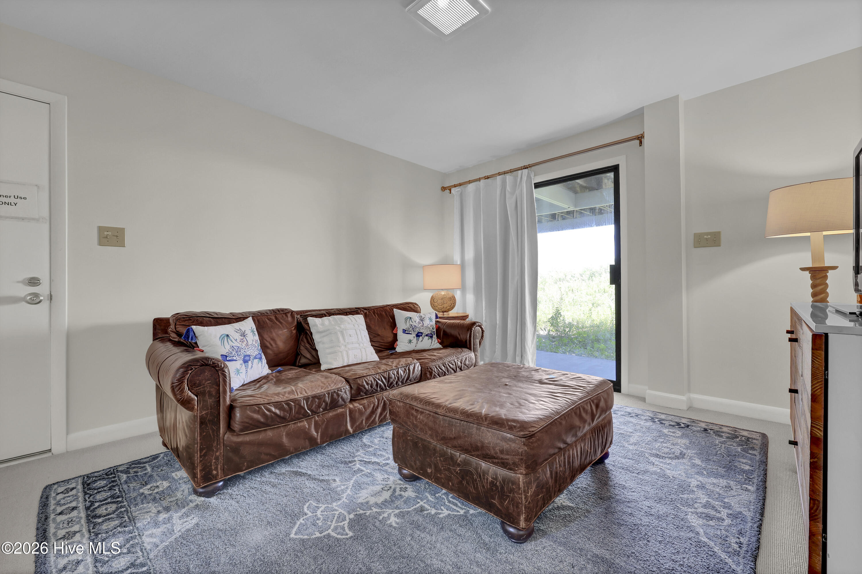Living room with brown leather sofa and ottoman beside a glass door opening to greenery.