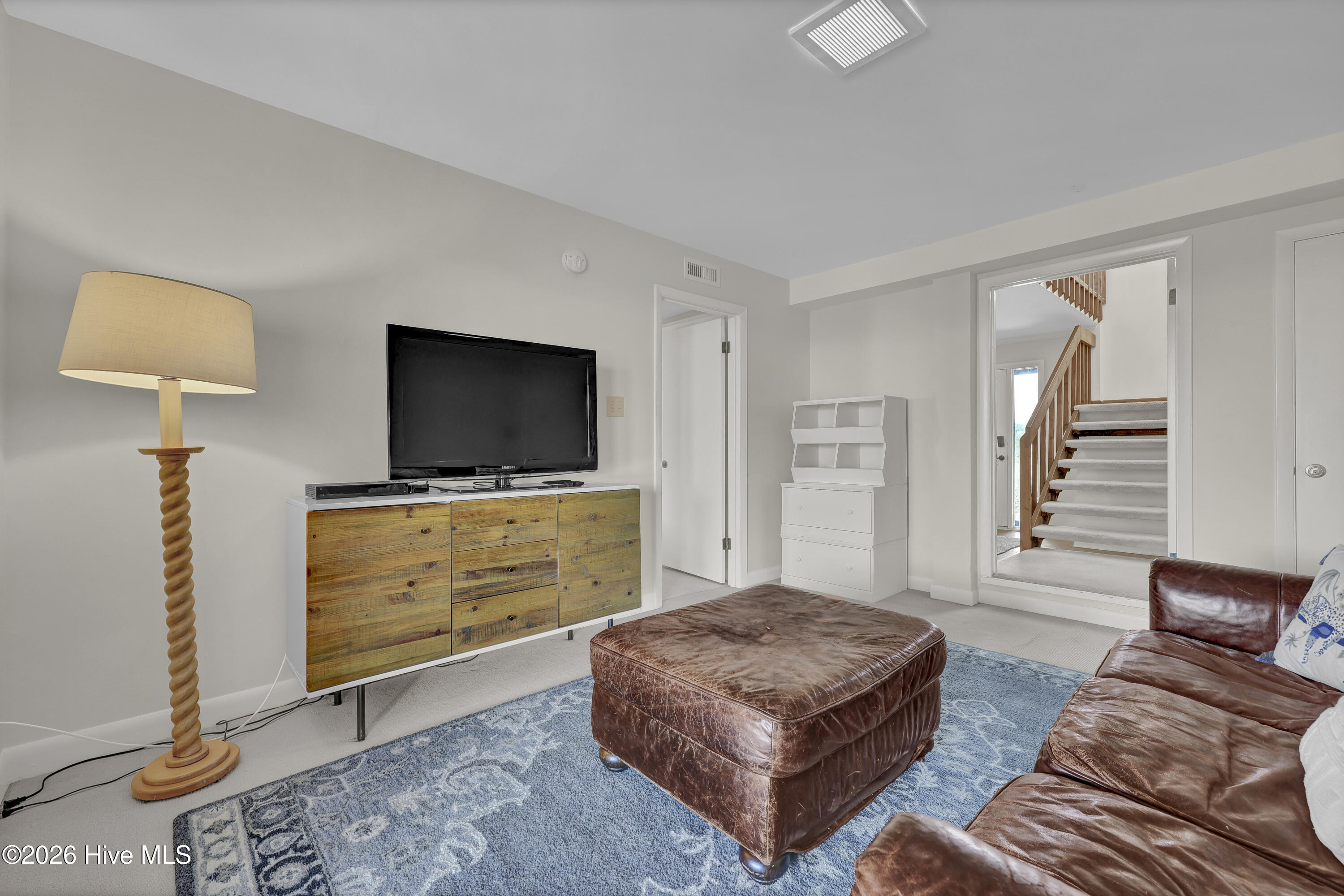Furnished living room with brown leather seating, a blue rug, and a staircase visible through the doorway.