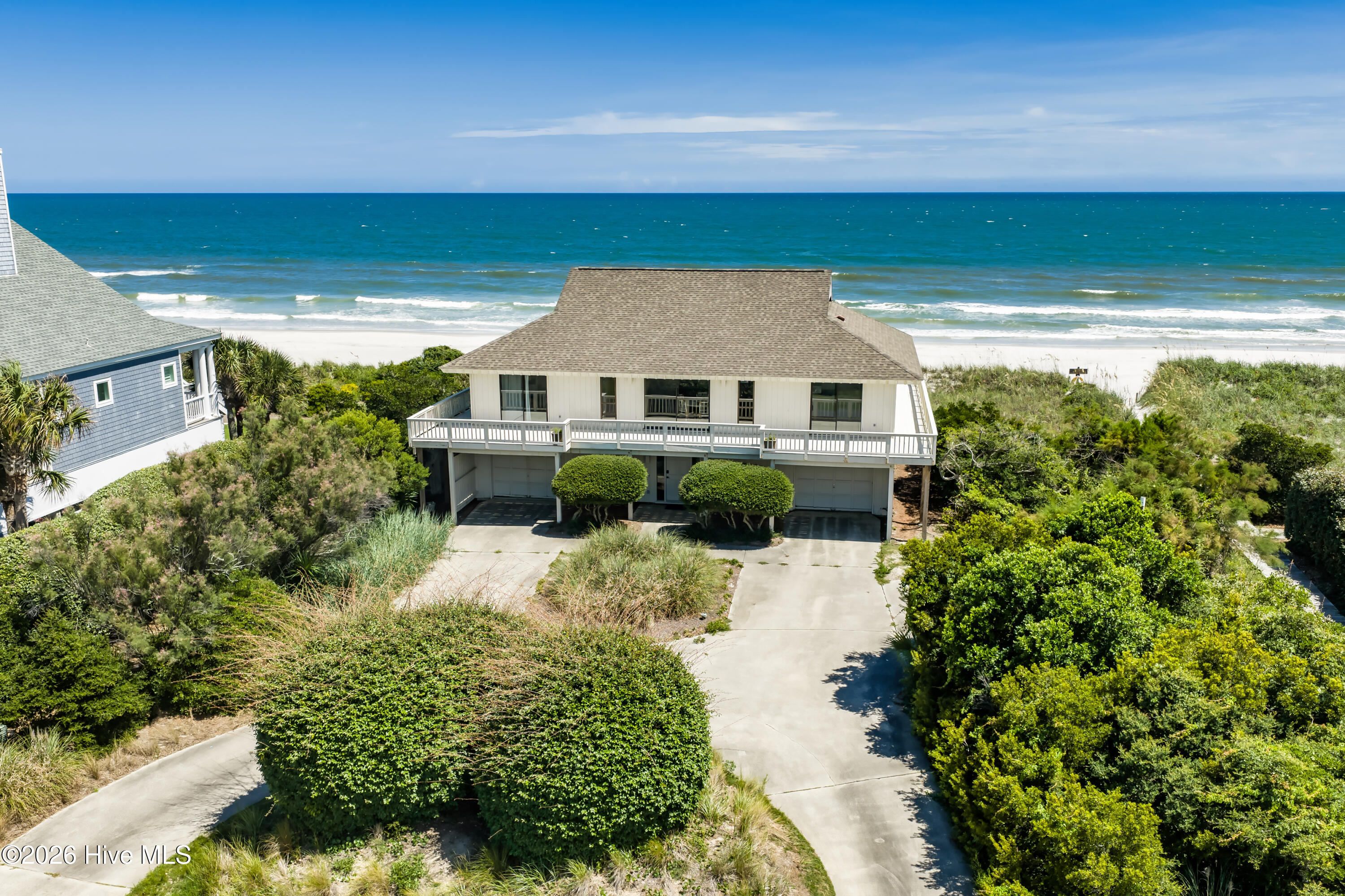 Raised beach house with a broad deck, landscaped grounds, and ocean waves behind it.