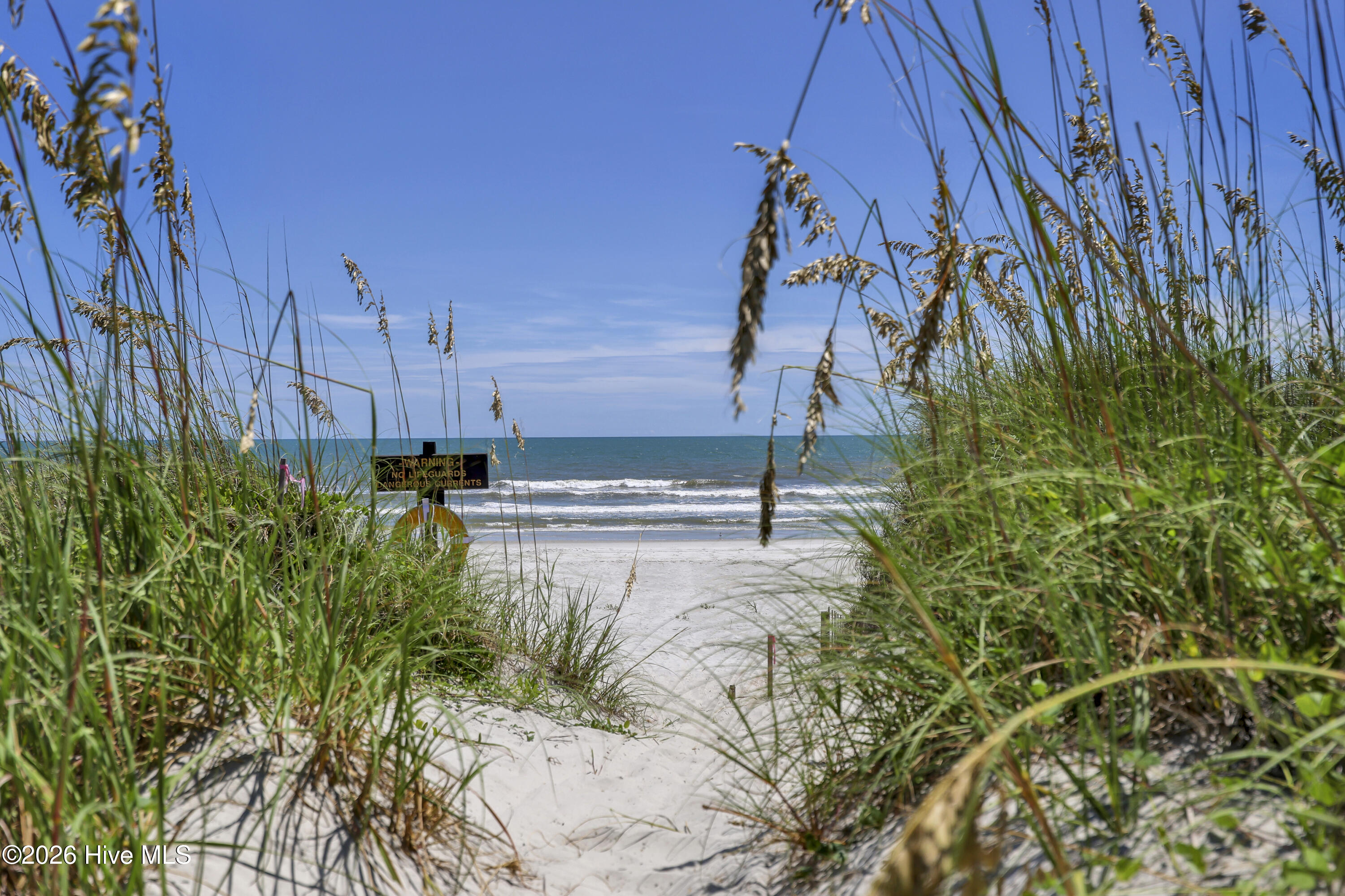 Sandy beach access path through dune grass leading to the ocean, with a warning sign.