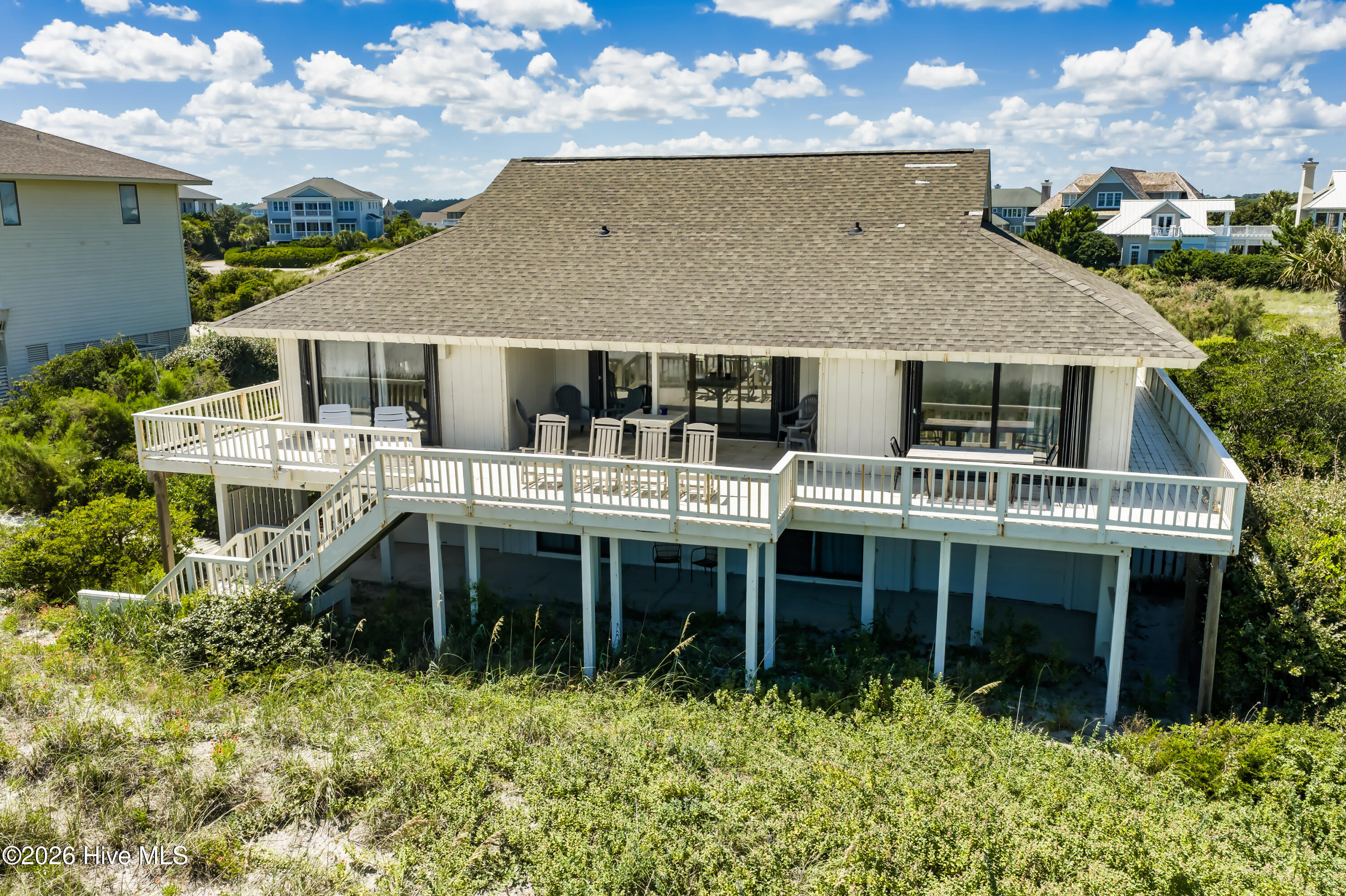 Raised coastal house with a broad wraparound deck amid dune grass and neighboring beach homes.