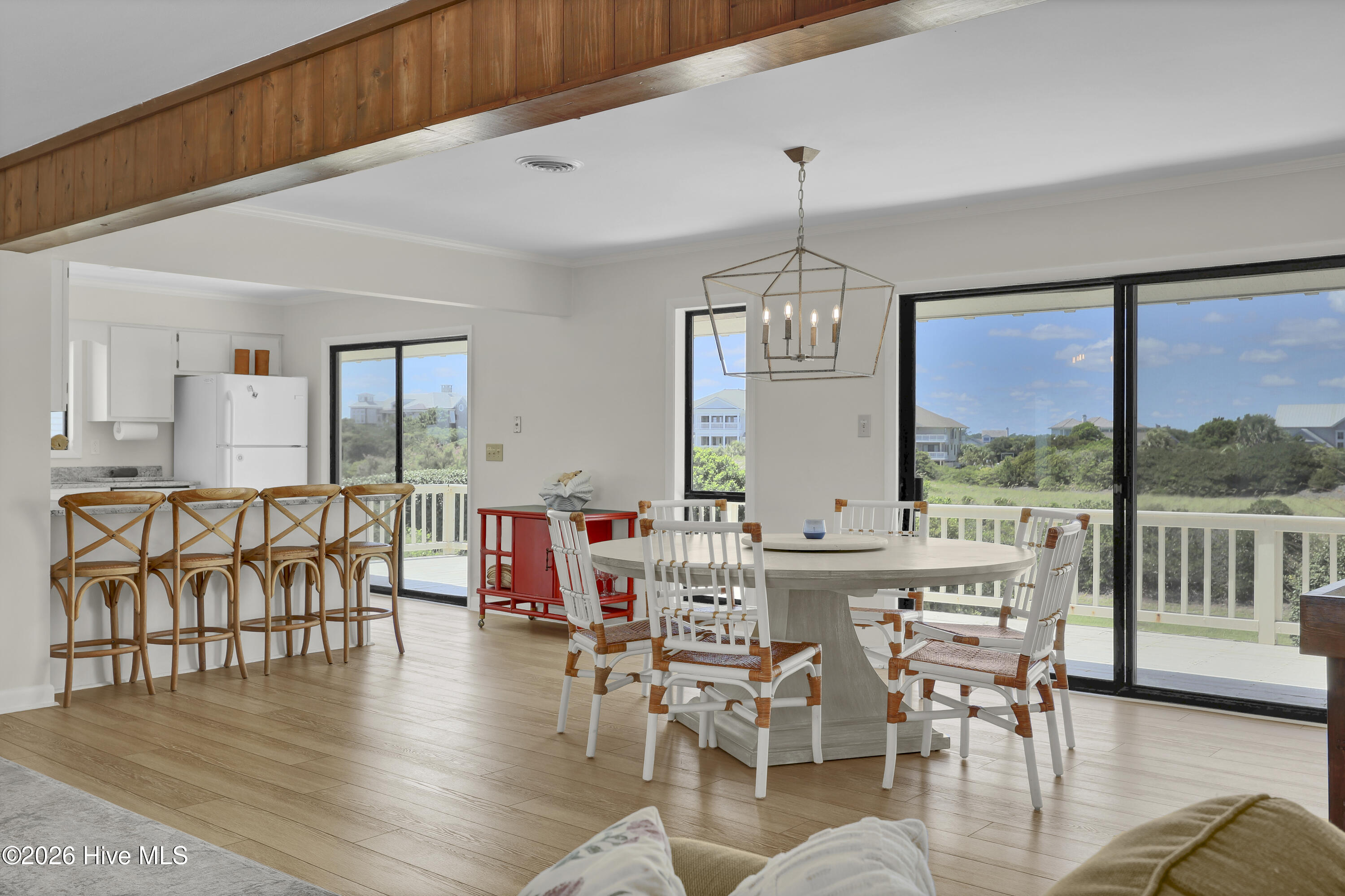 Open dining area with a round table, woven chairs, wood ceiling beam, and large windows overlooking coastal greenery.
