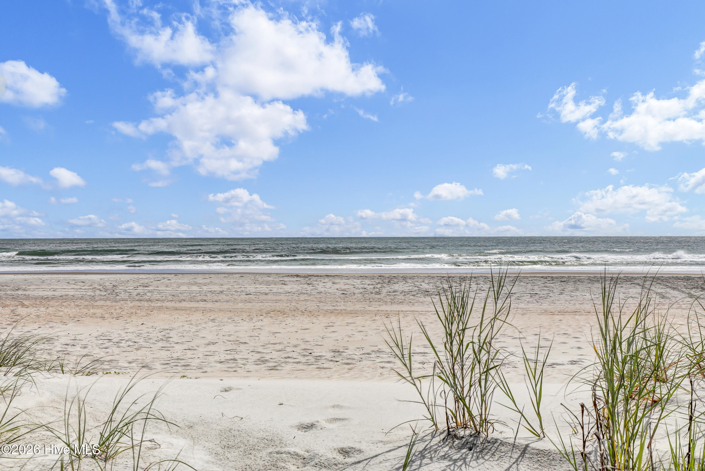 Sandy beach and ocean waves beneath a cloud-filled blue sky, framed by dune grass.
