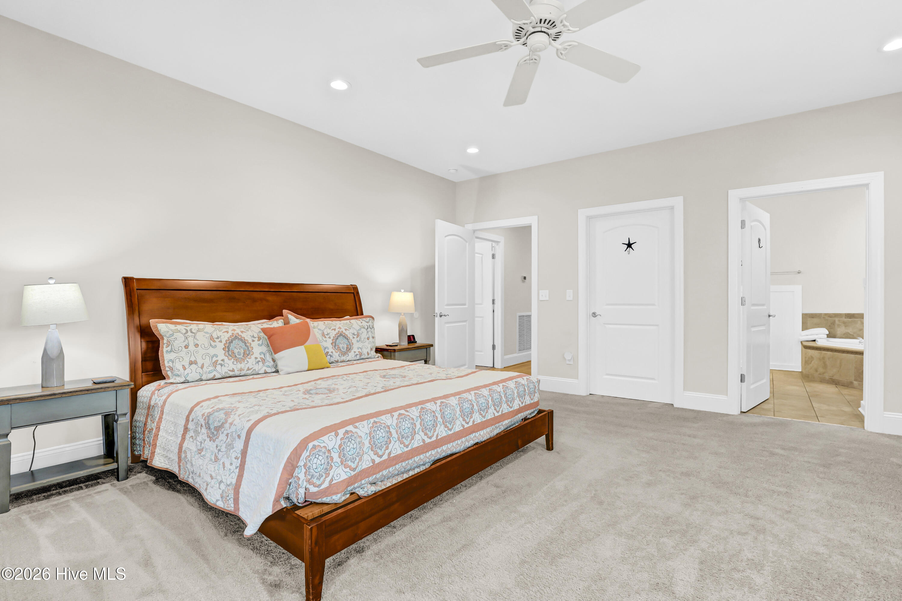 Furnished bedroom with a wooden bed, patterned quilt, ceiling fan, and doorway to a tiled bathroom.