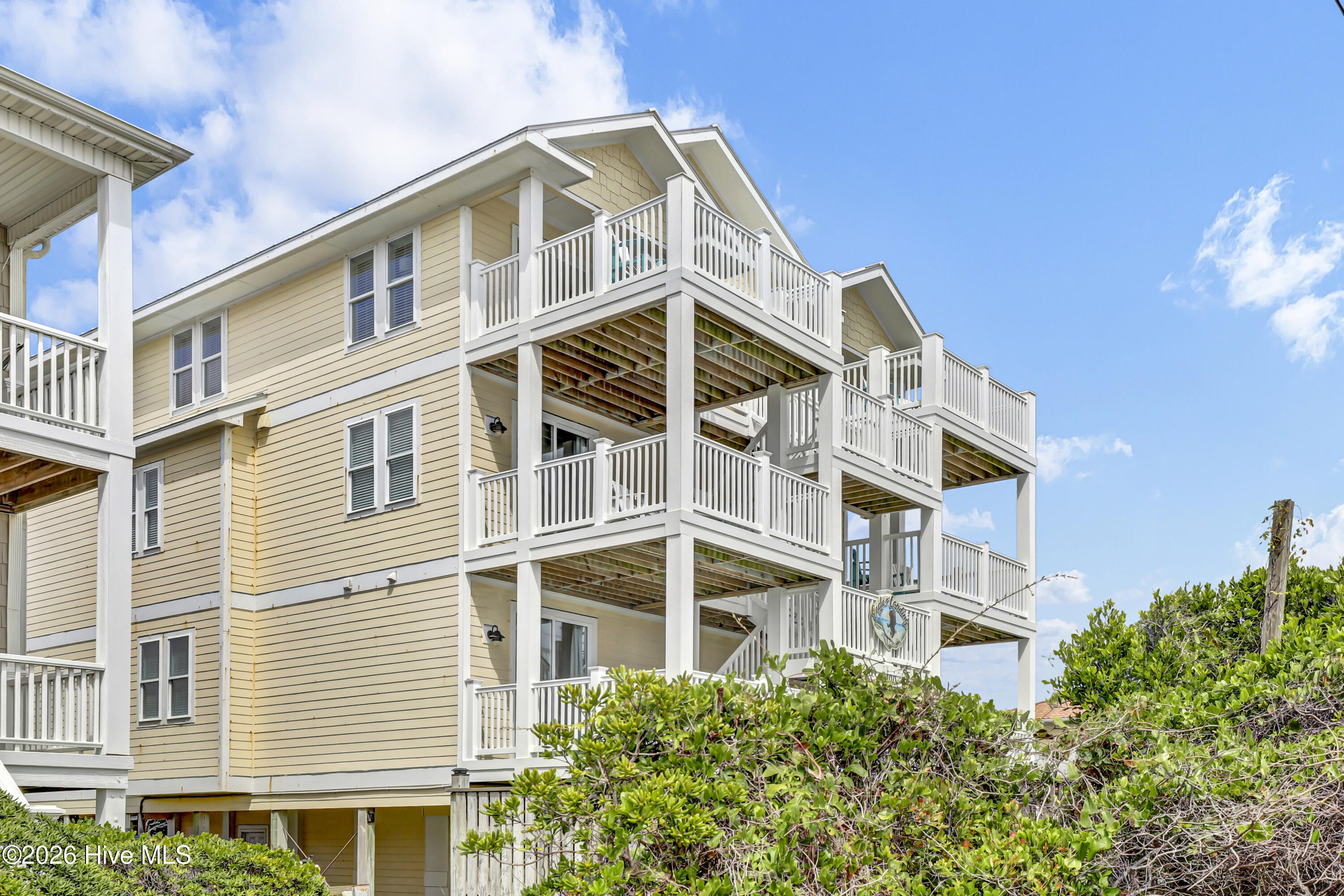 Pale yellow coastal home with stacked white decks, elevated above dense shrubs.