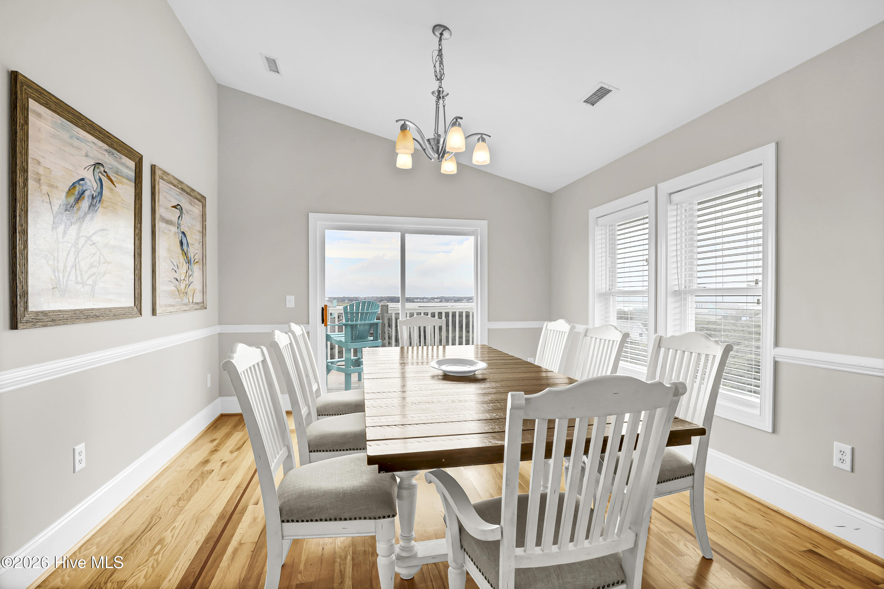 Dining room with a wooden table, white chairs, and windows overlooking water and a deck.
