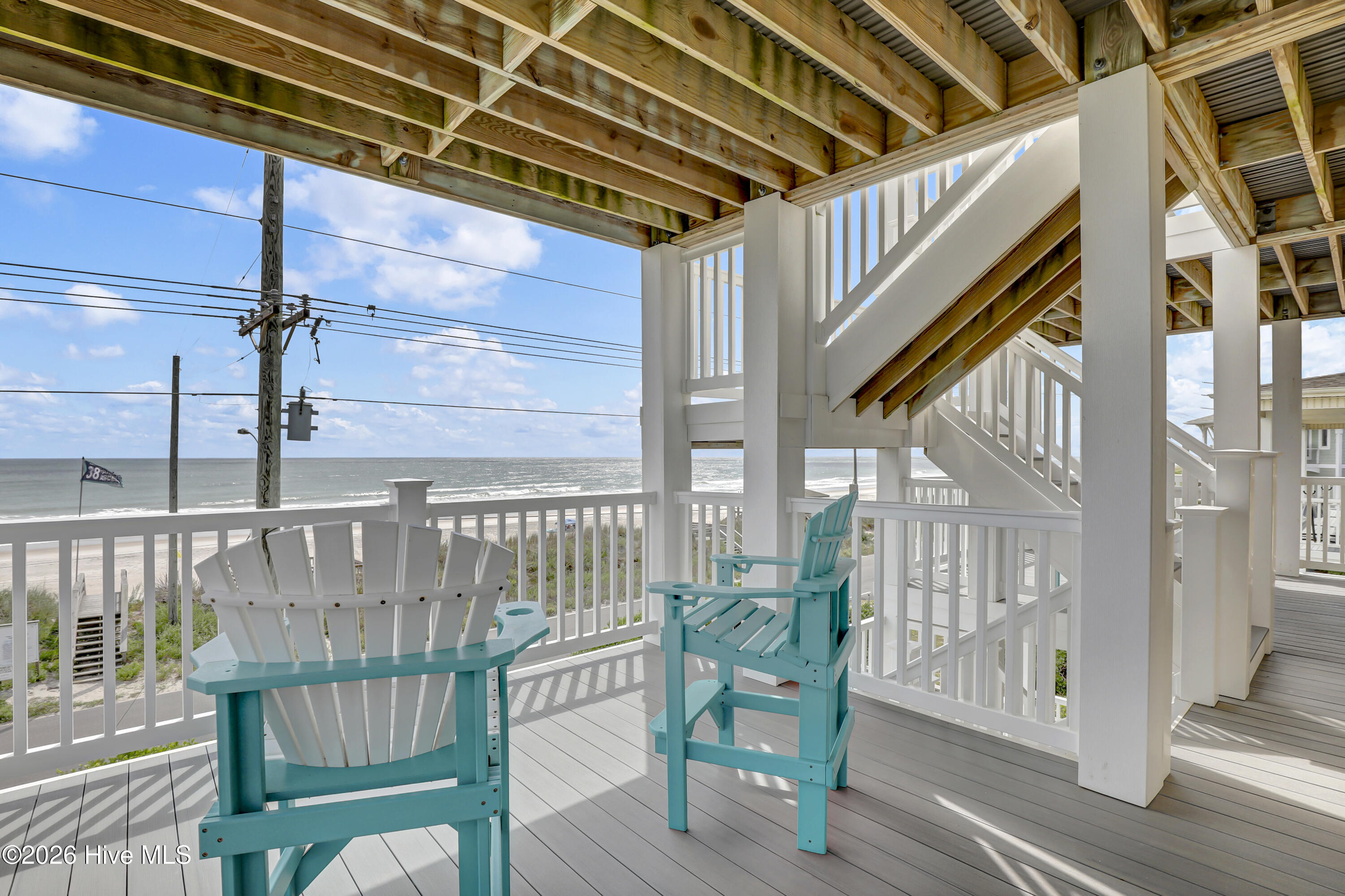 Covered oceanfront deck with turquoise Adirondack chairs, white railings, and beach and ocean views.
