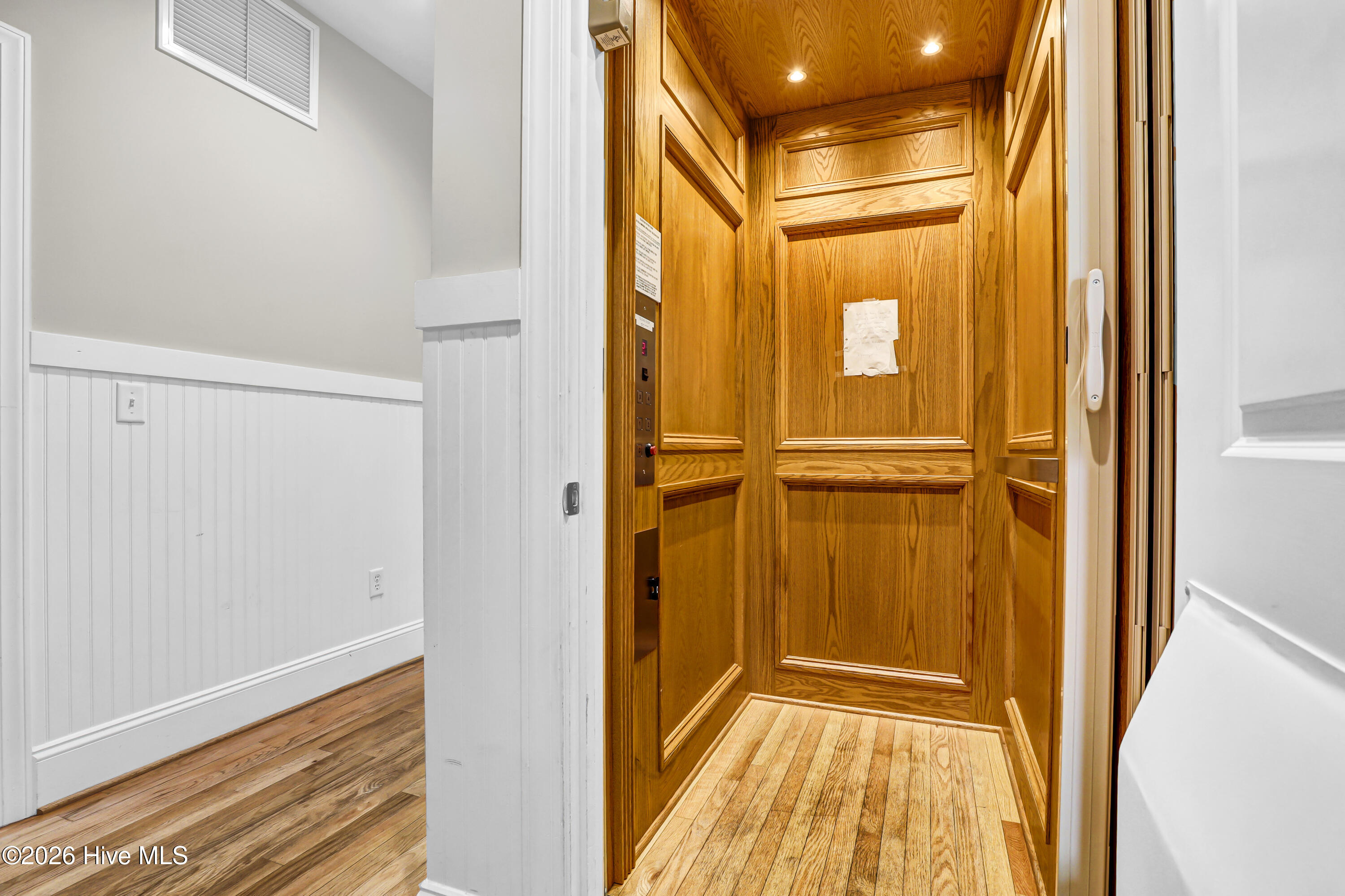 Wood-paneled residential elevator with recessed lights and control buttons, opening onto a white-trimmed hallway.