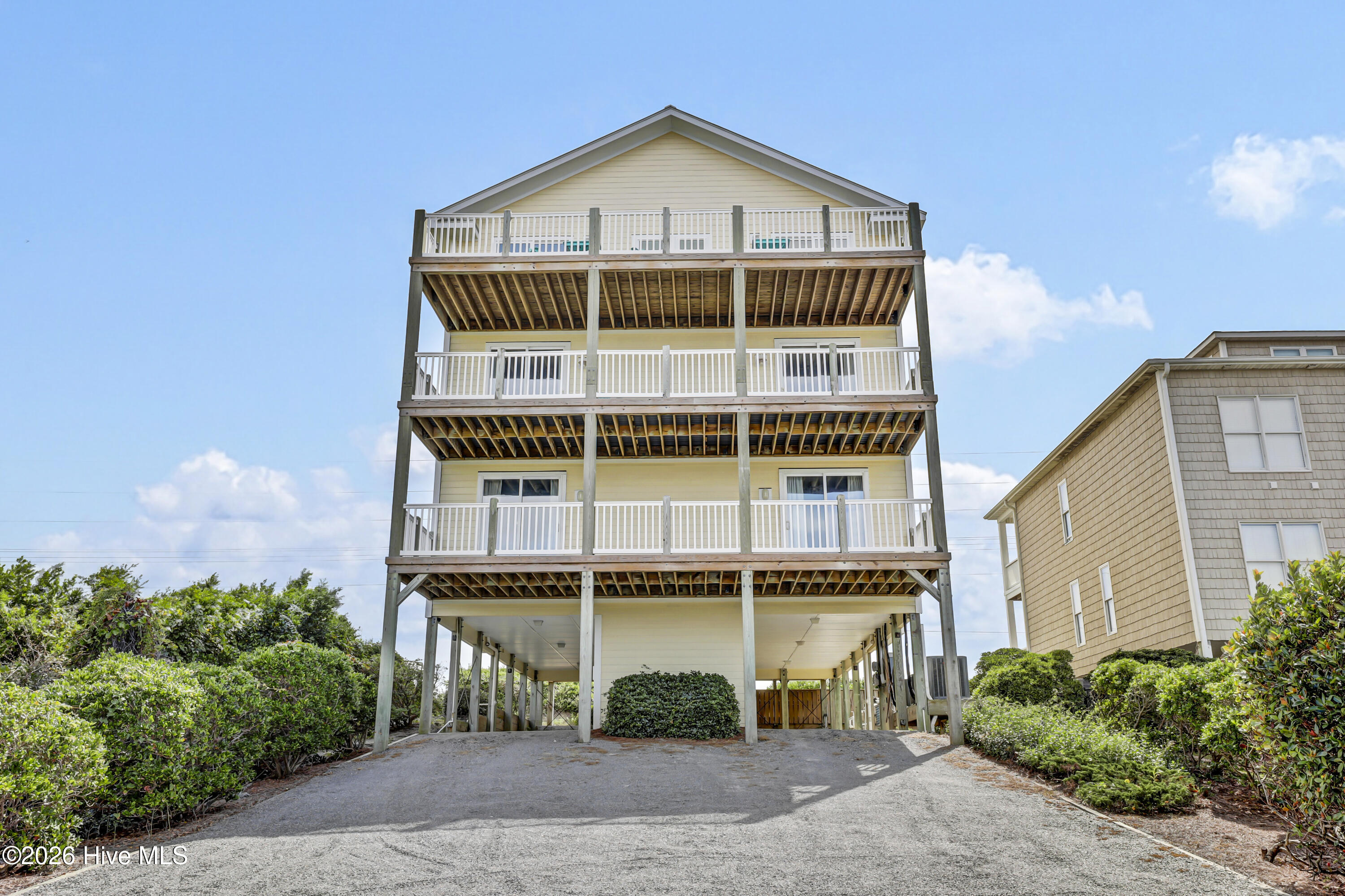 Elevated pale yellow coastal house with three stacked white-railed balconies and a gravel driveway.