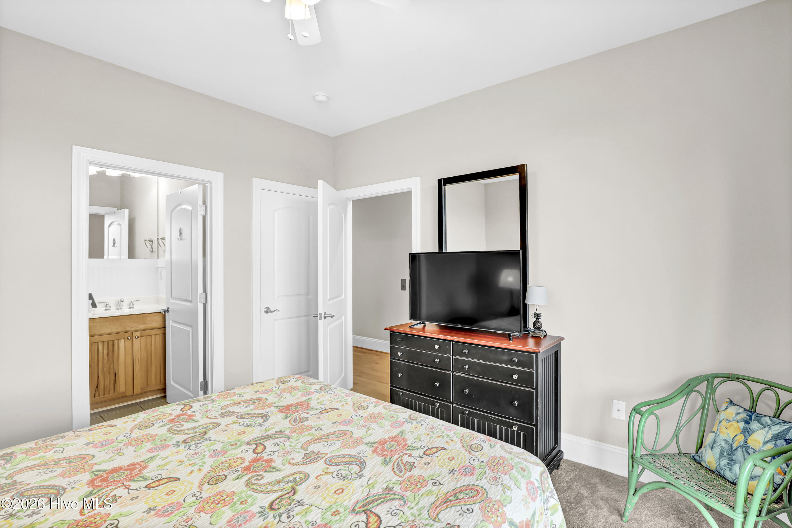 Bedroom with a colorful patterned bedspread, black dresser and TV, green wicker chair, and adjoining bathroom.
