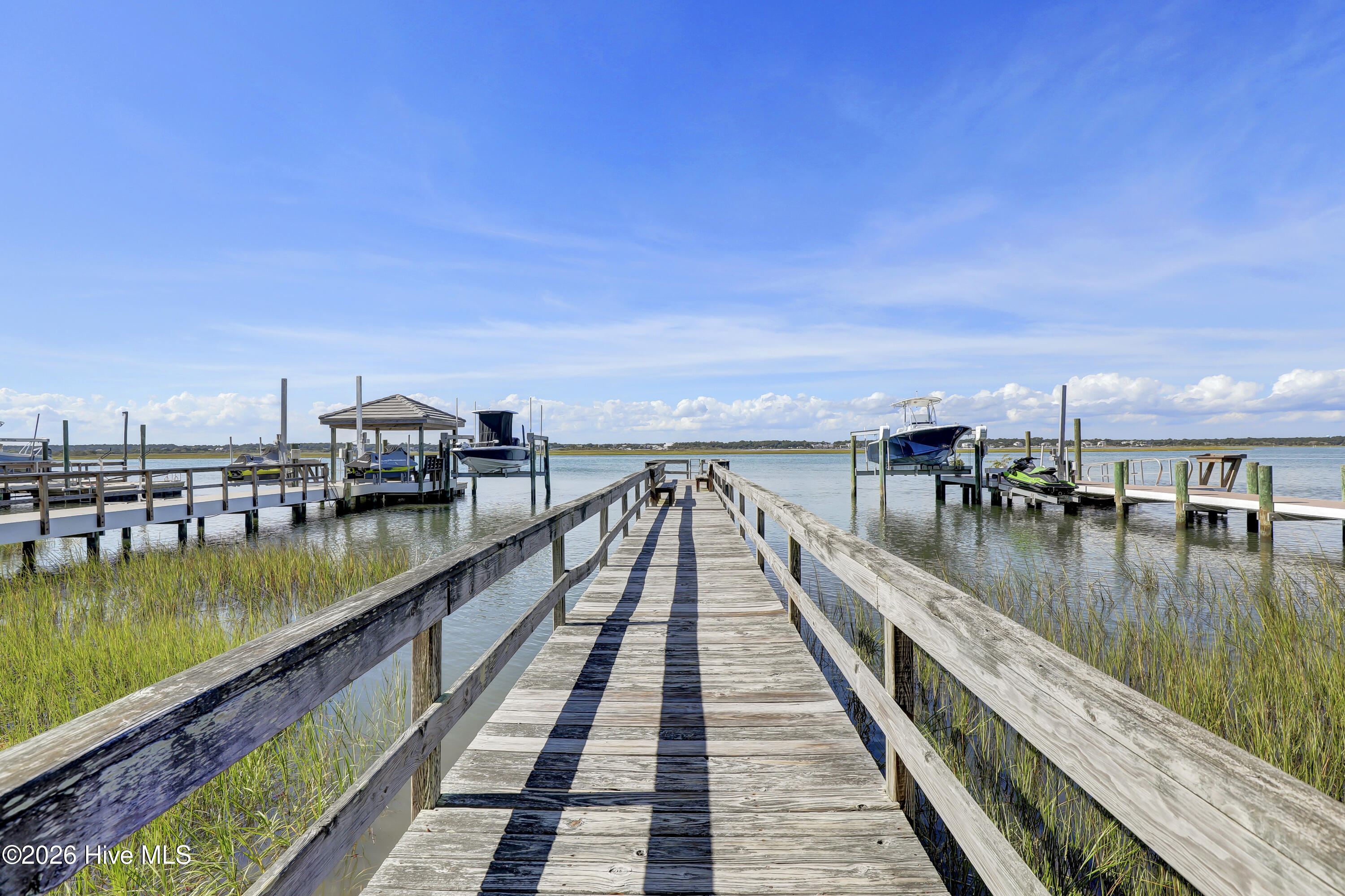 Wooden dock walkway through marsh grass toward boat lifts and coastal water.