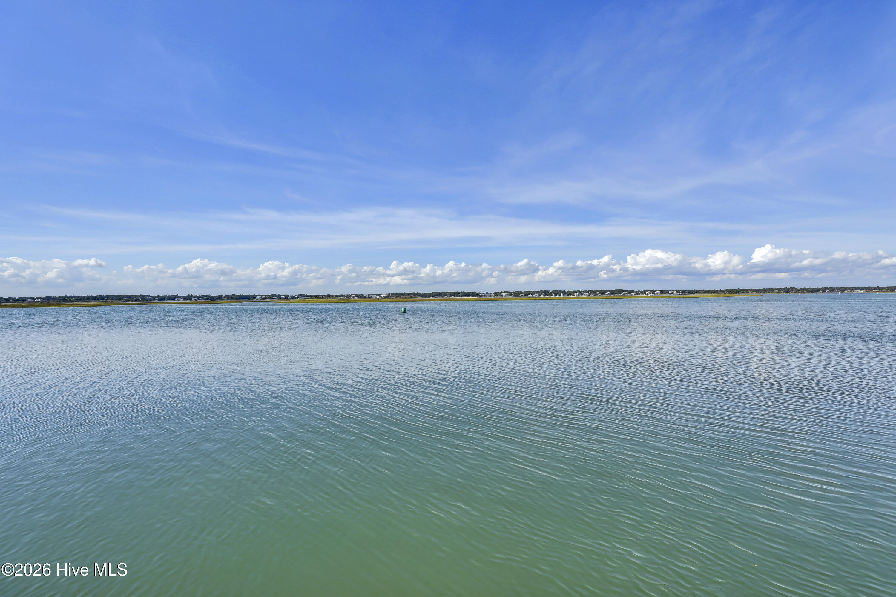 Wide view across calm water toward a distant shoreline and homes beneath a cloud-filled blue sky.
