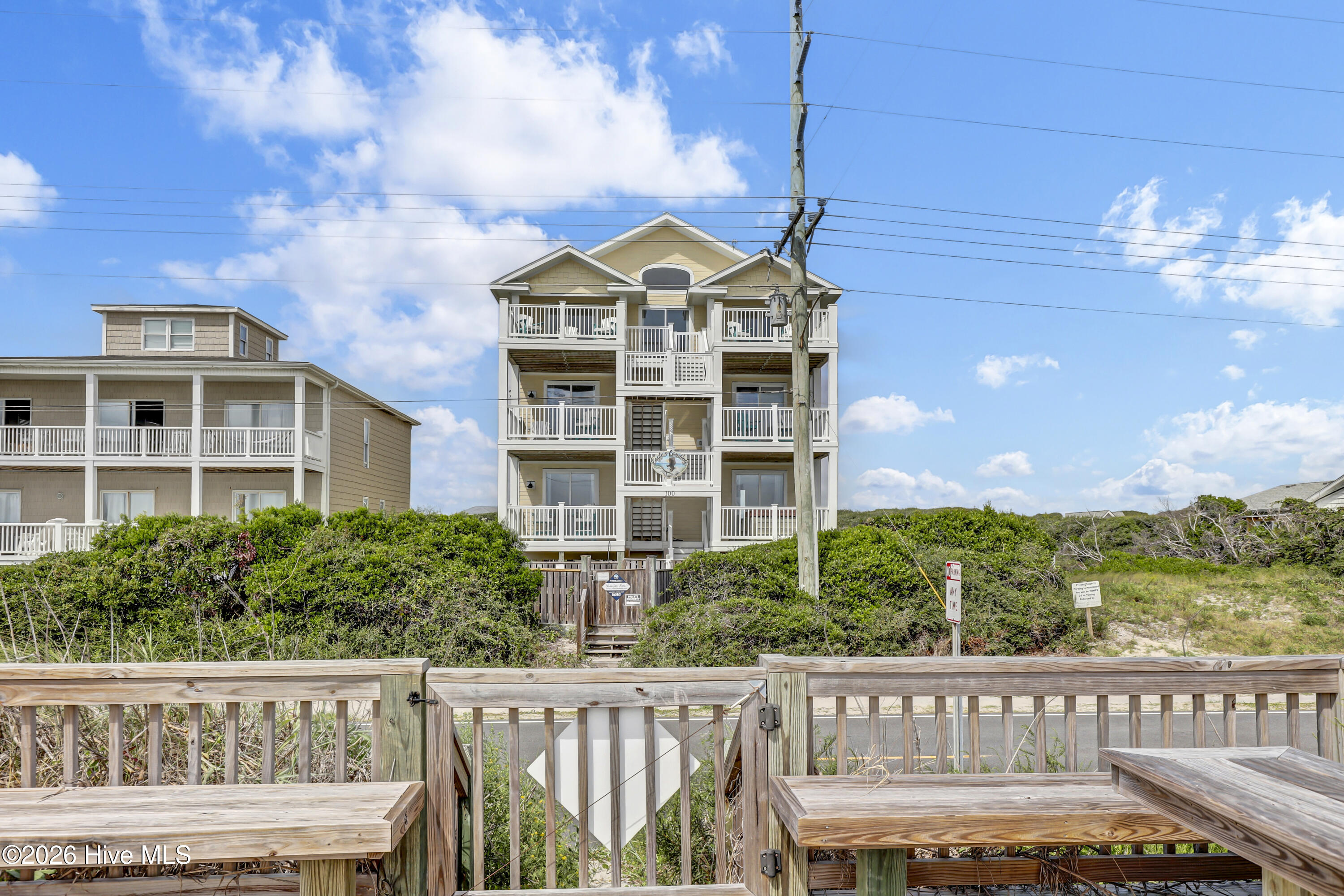 Coastal house with stacked white-railed balconies behind dune shrubs and a wooden boardwalk.