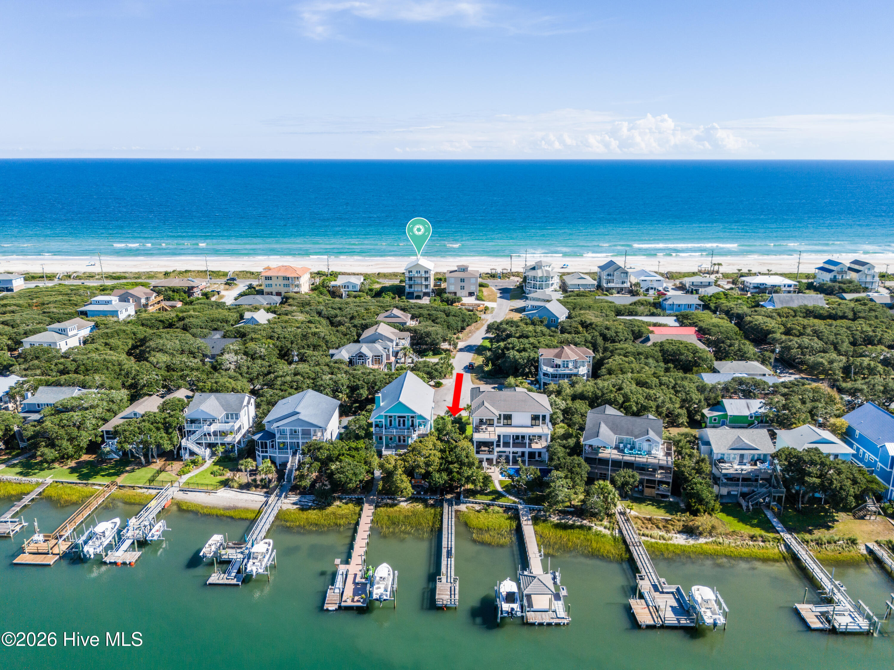 Aerial view of coastal homes between the ocean and a dock-lined waterway, with the featured house marked by an arrow.
