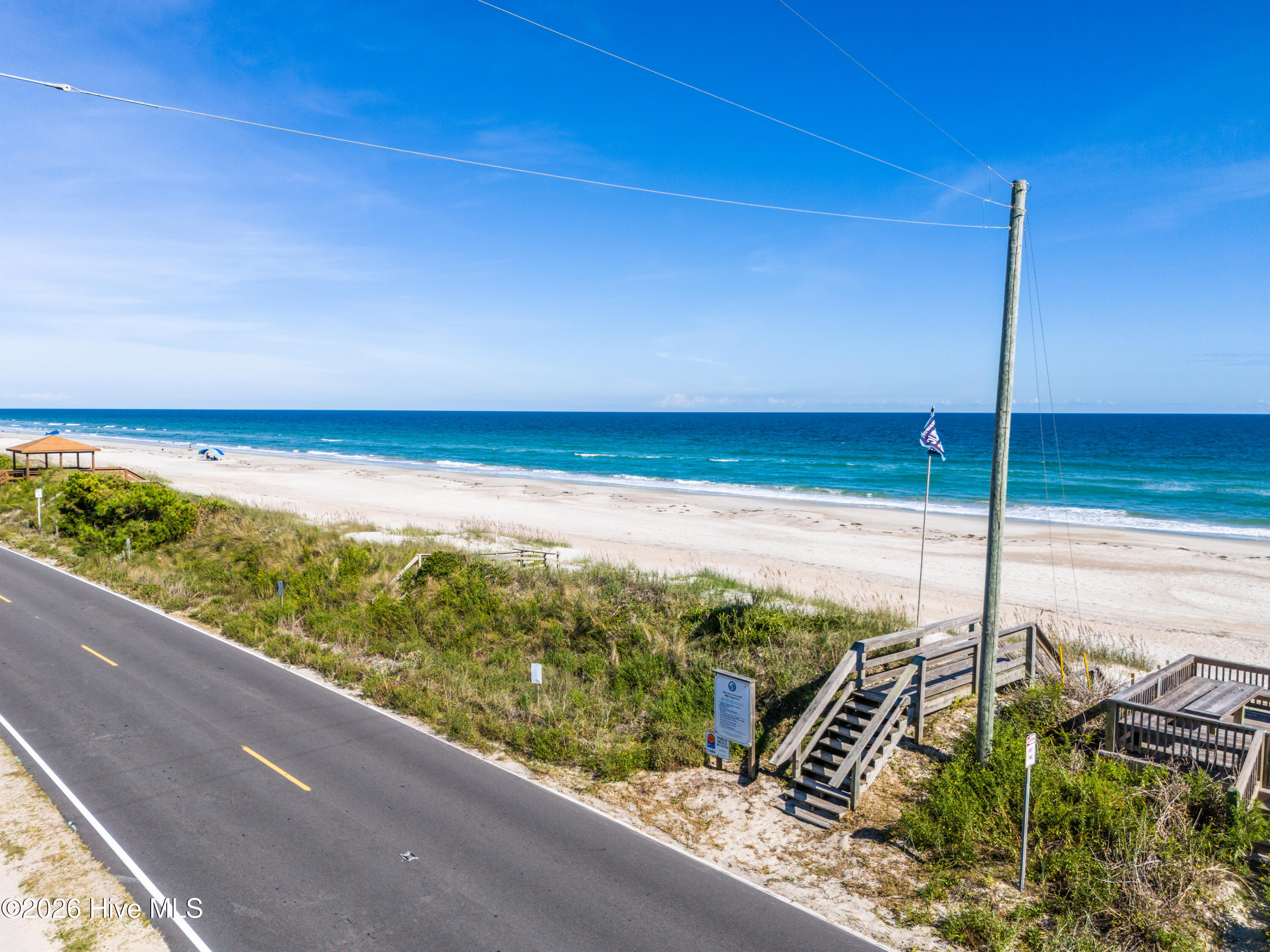 Sandy beach and ocean beyond dune grass, with wooden beach-access stairs beside a coastal road.