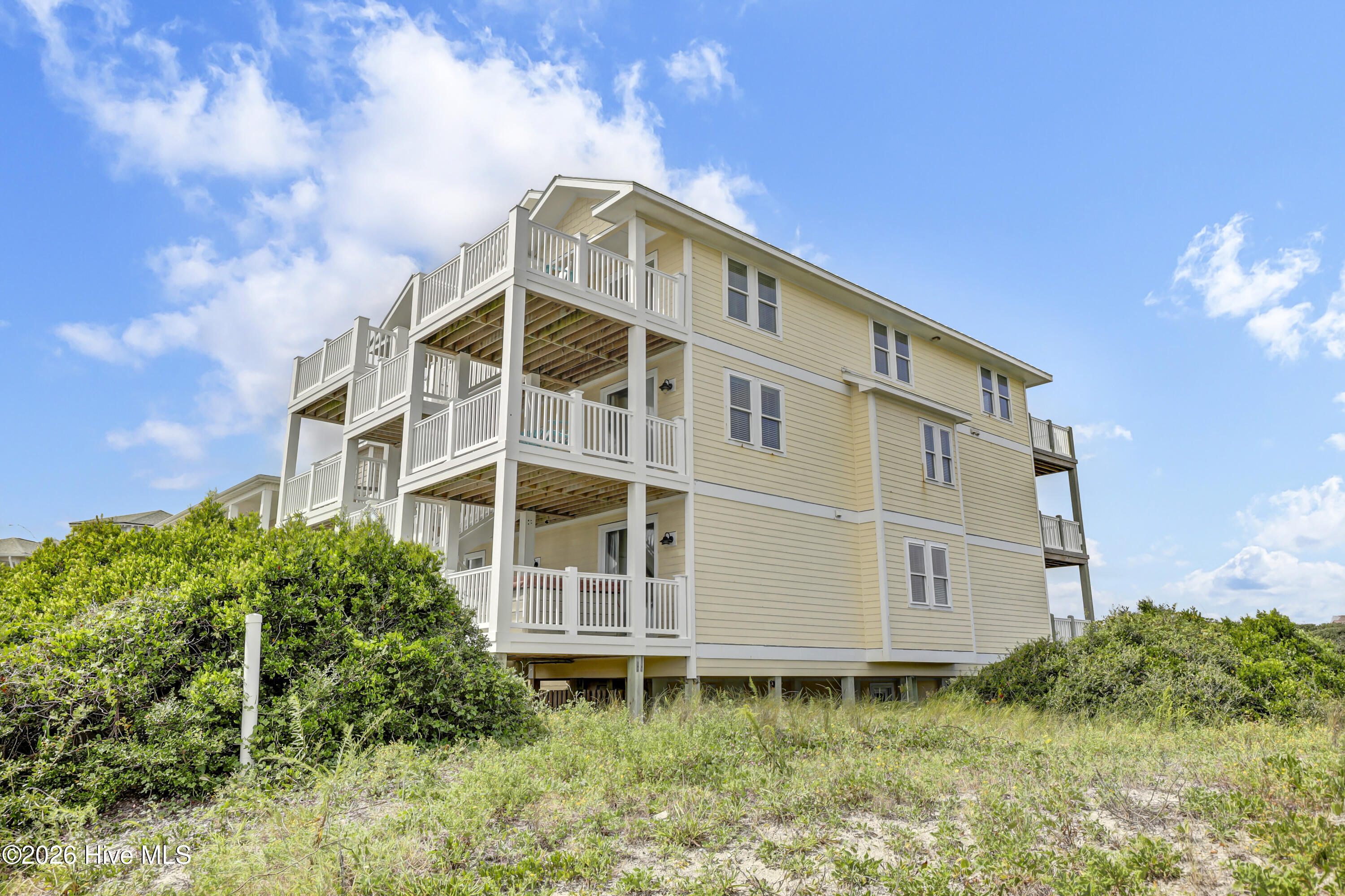Raised pale yellow coastal house with white multi-level decks amid dune grasses and shrubs.