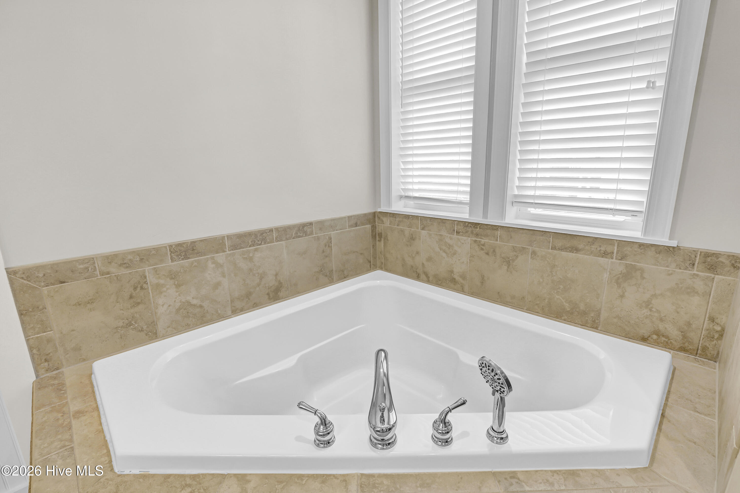 Jetted bathtub with chrome fixtures, tan tile surround, and windows with white blinds.
