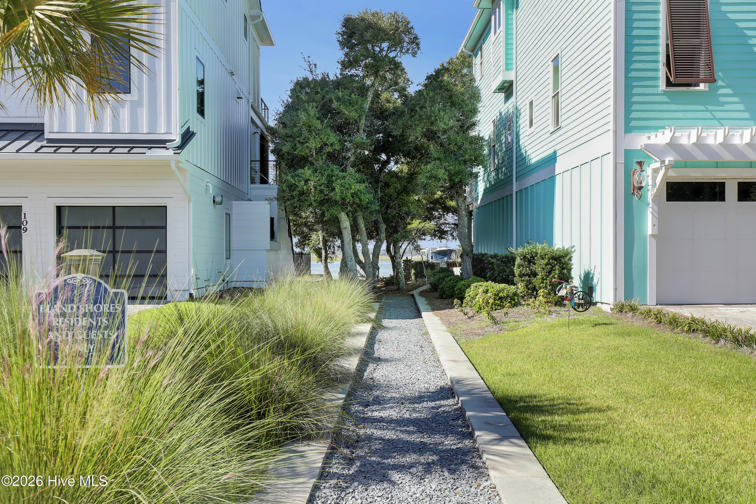 Gravel path between coastal homes, with ornamental grasses and a glimpse of water beyond the trees.
