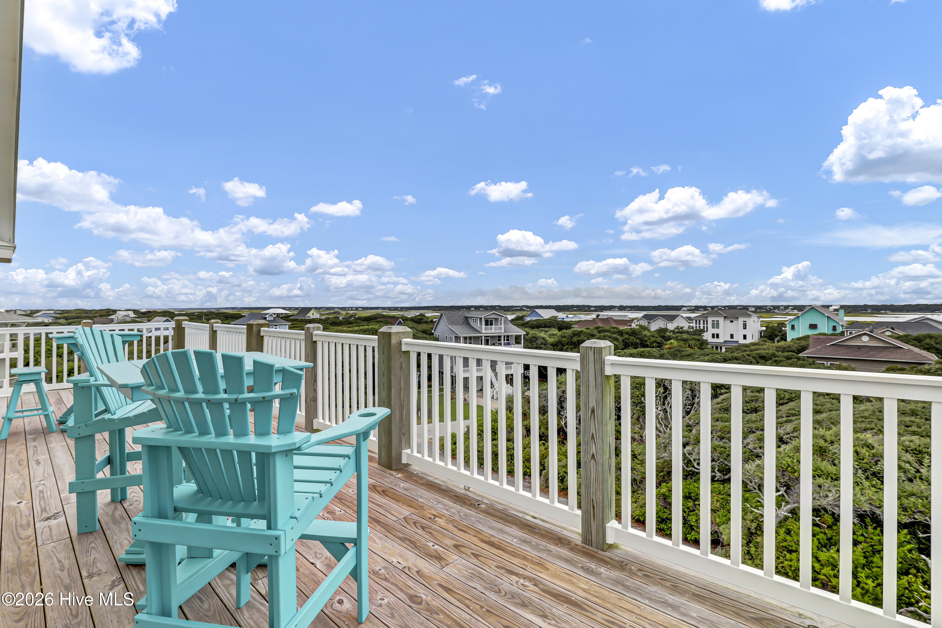 Raised wooden deck with turquoise Adirondack chairs overlooking coastal vegetation and neighboring homes.