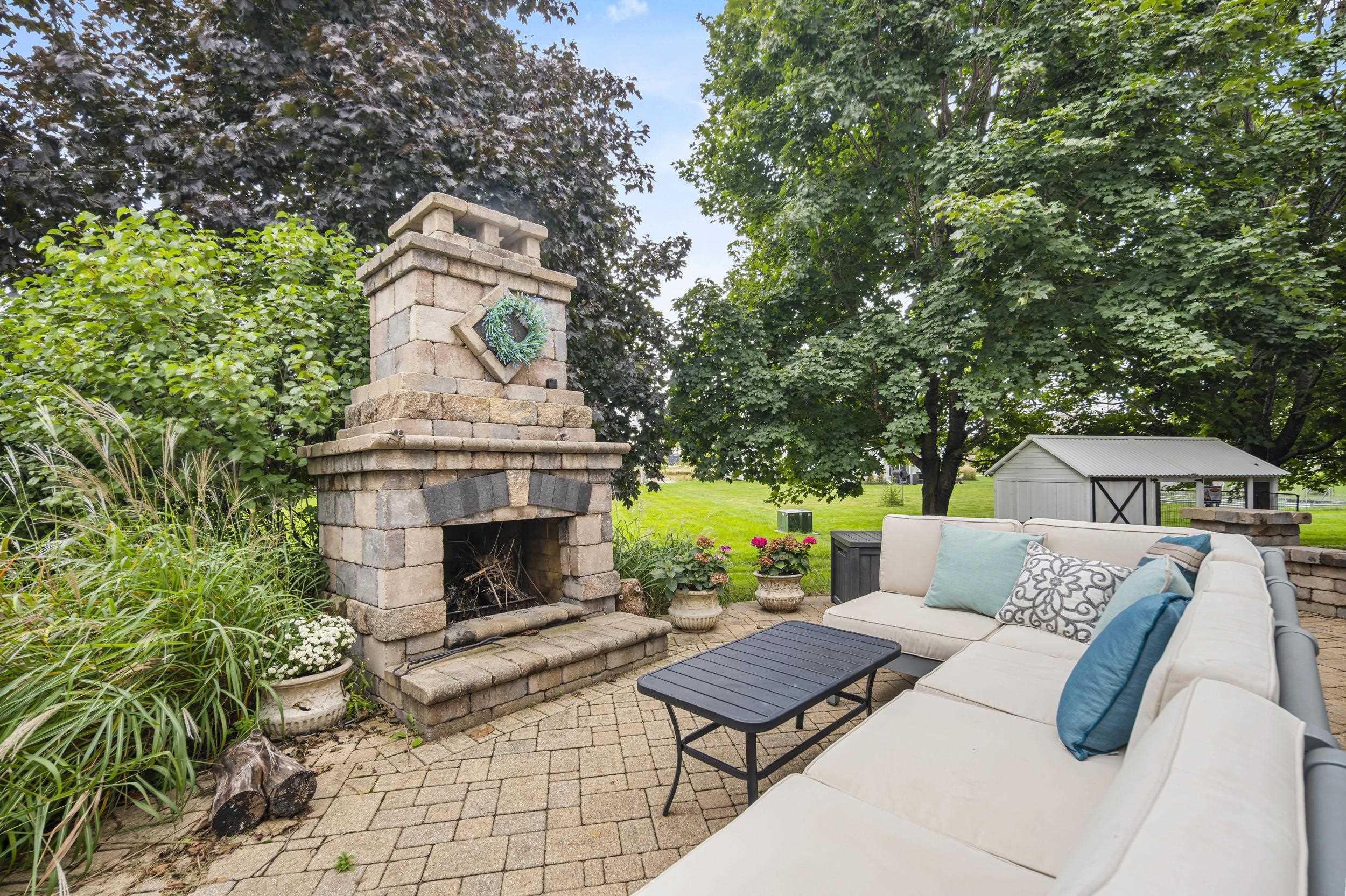 Backyard patio with a stone fireplace, cushioned sectional, pavers, and a leafy landscaped yard.