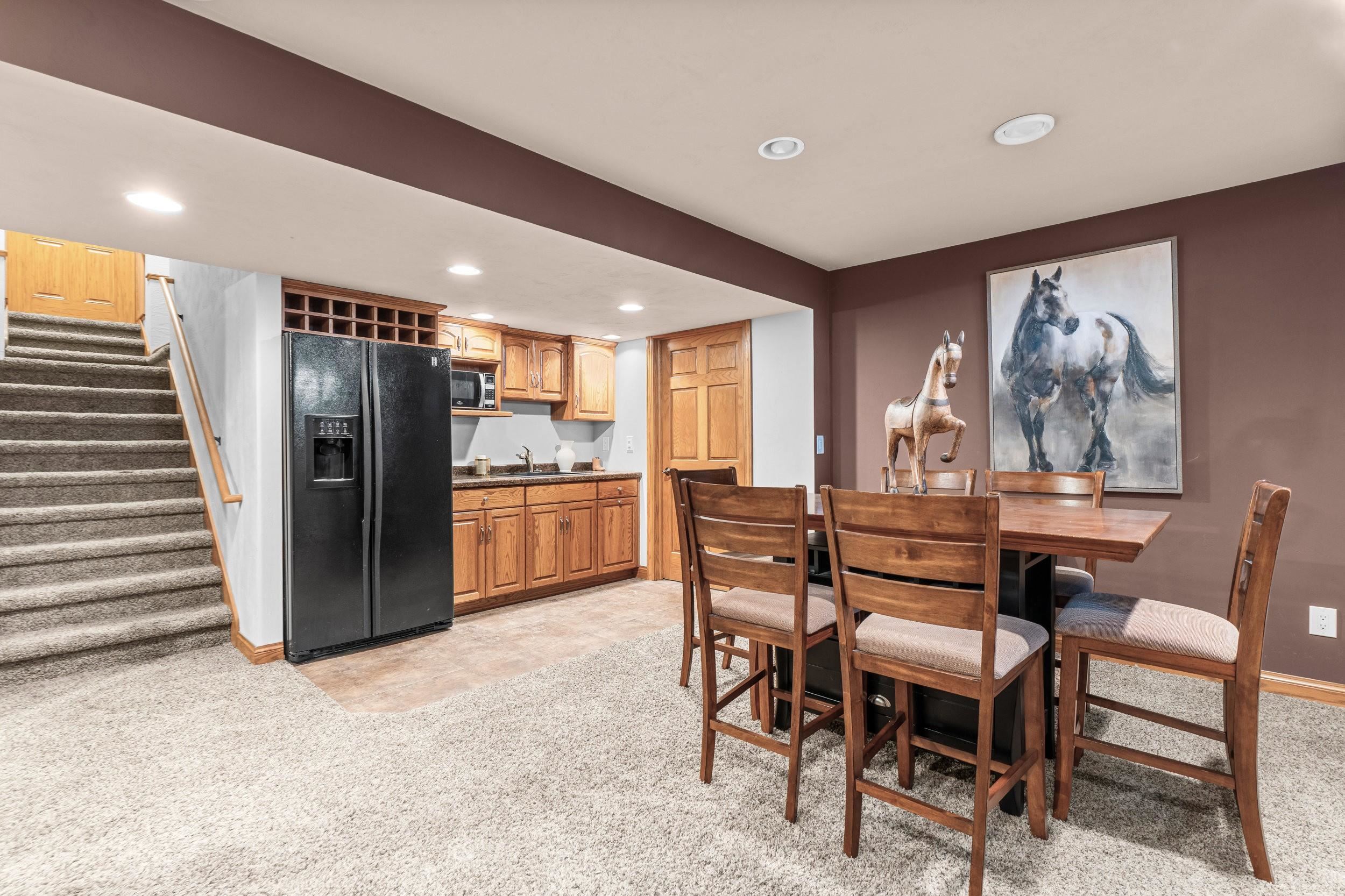 Basement dining area with an oak-cabinet kitchenette, black refrigerator, and carpeted stairs.