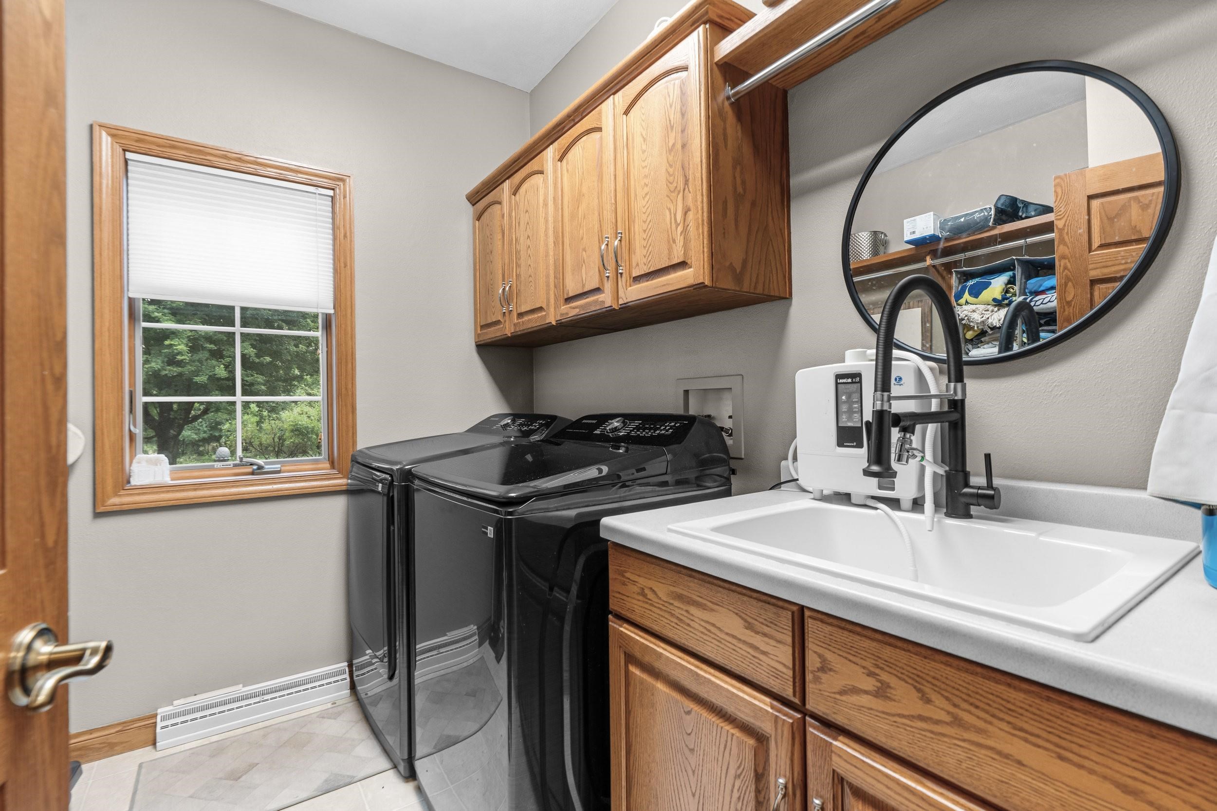 Laundry room with black washer and dryer, wood cabinets, and a utility sink beside a window.