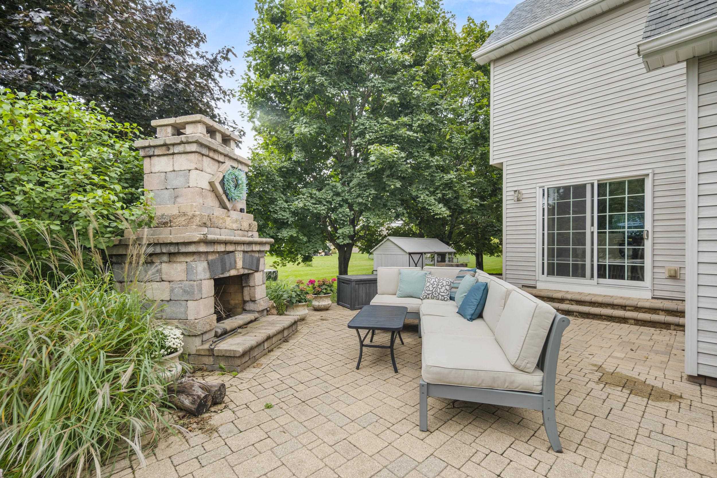 Backyard patio with a stone fireplace, cushioned sectional, and leafy trees.