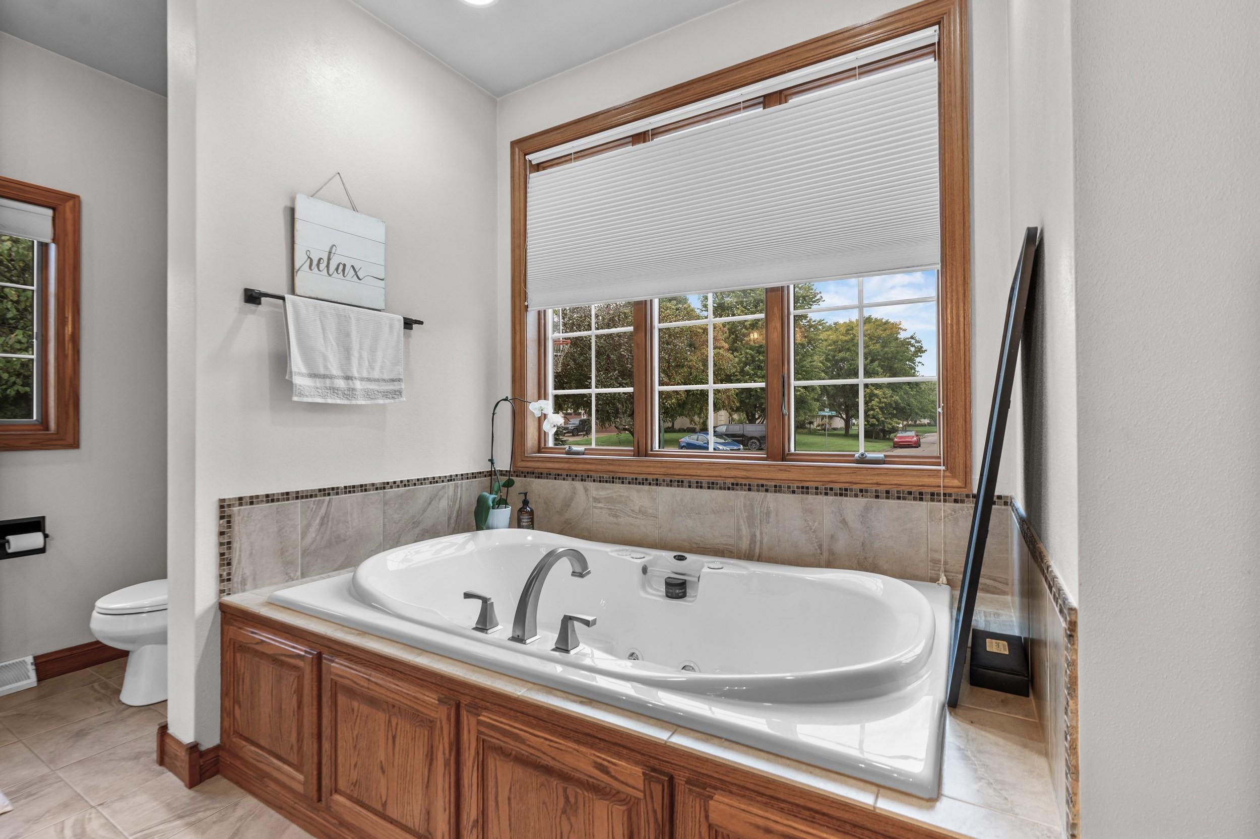 Bathroom with a jetted soaking tub beneath a wide window overlooking a tree-lined street.