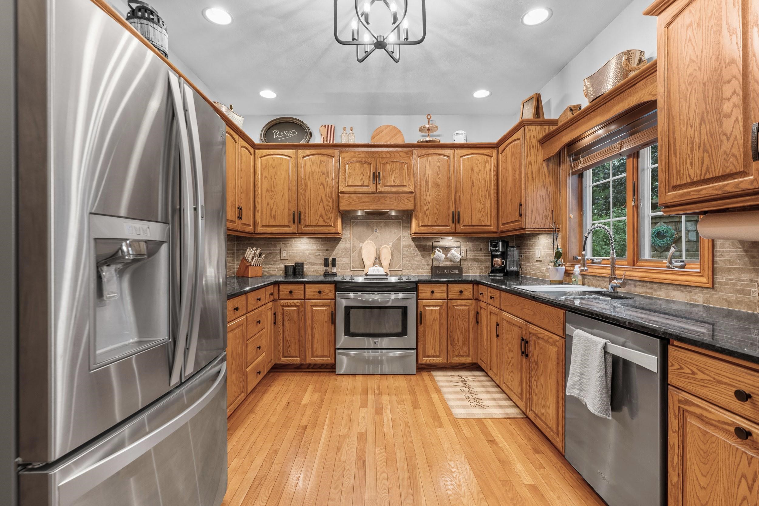 Kitchen with oak cabinetry, stainless steel appliances, dark counters, and a large window over the sink.