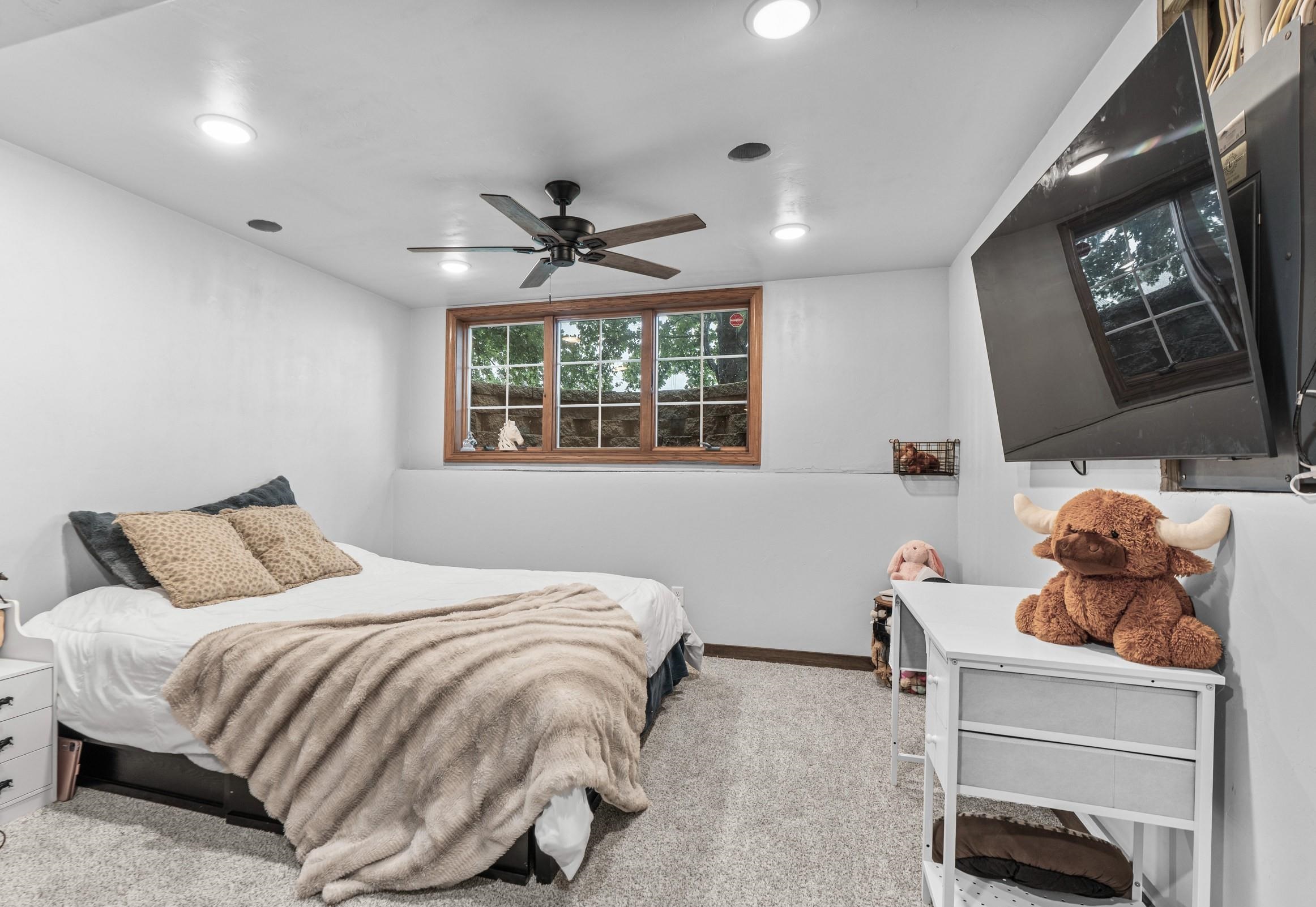 Furnished bedroom with a beige throw on the bed, wood-trimmed window, ceiling fan, and wall-mounted television.