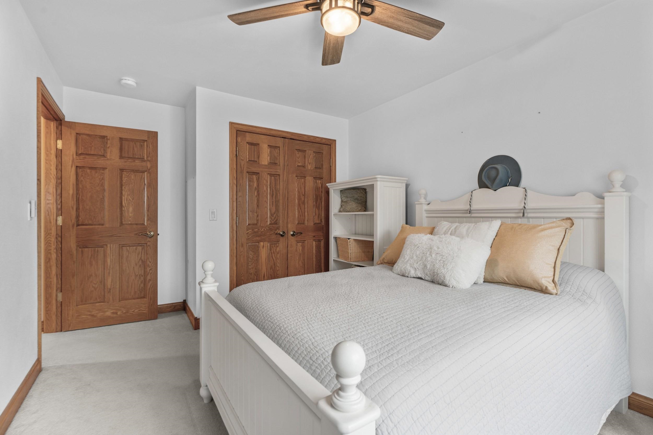 Staged bedroom with a white bed, gold accent pillows, wood doors, and a lit ceiling fan.