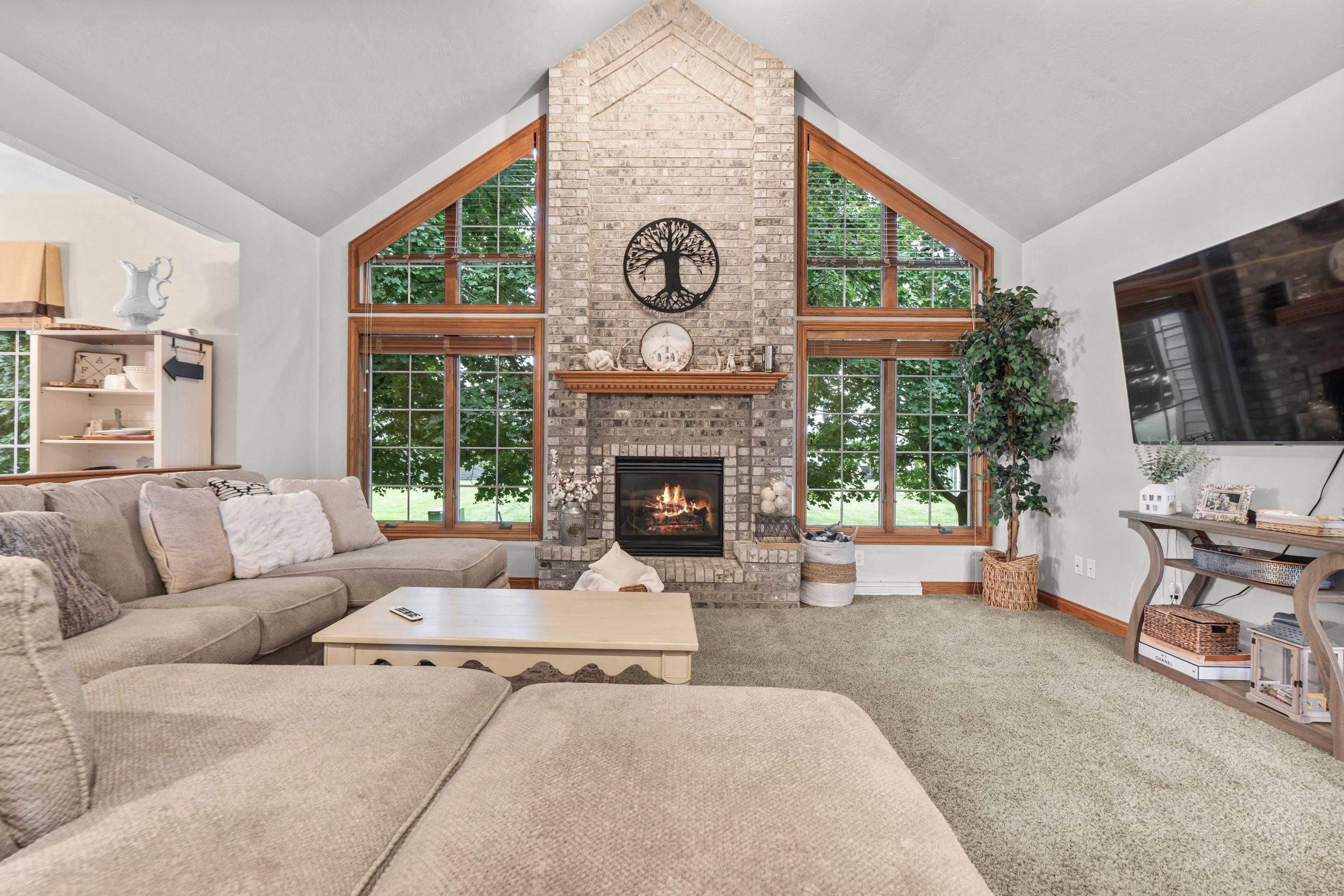Living room with a tall brick fireplace, vaulted ceiling, and large wood-trimmed windows.
