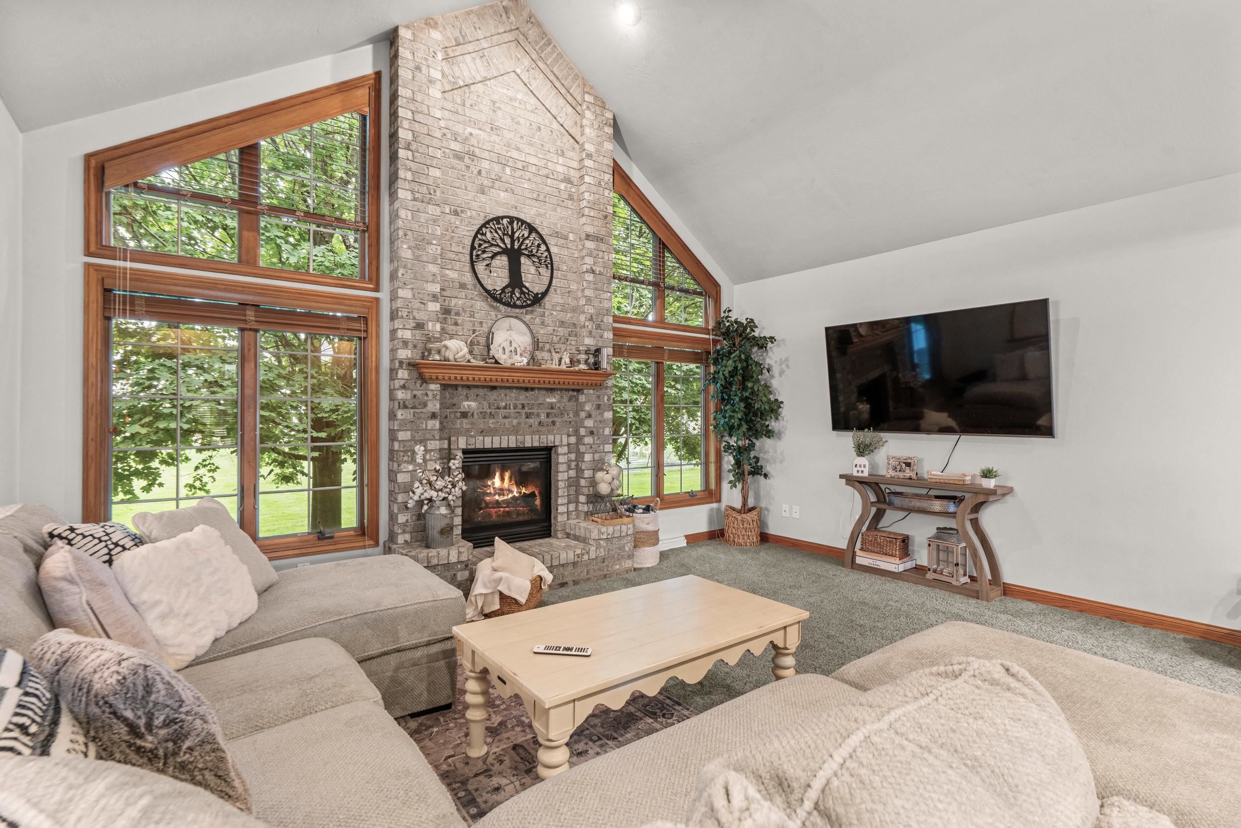 Living room with a tall brick fireplace, vaulted ceiling, and large windows overlooking trees.