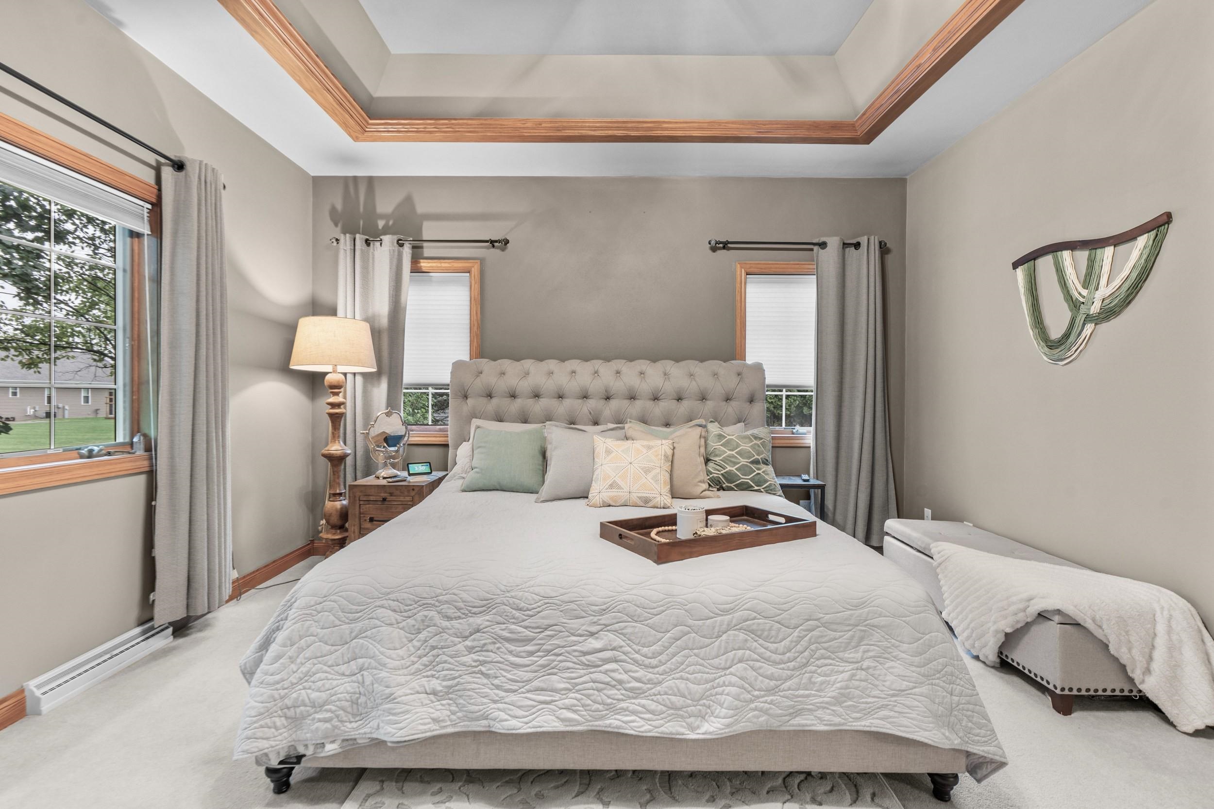 Staged bedroom with a tufted headboard, wood-trimmed windows, and a tray ceiling.