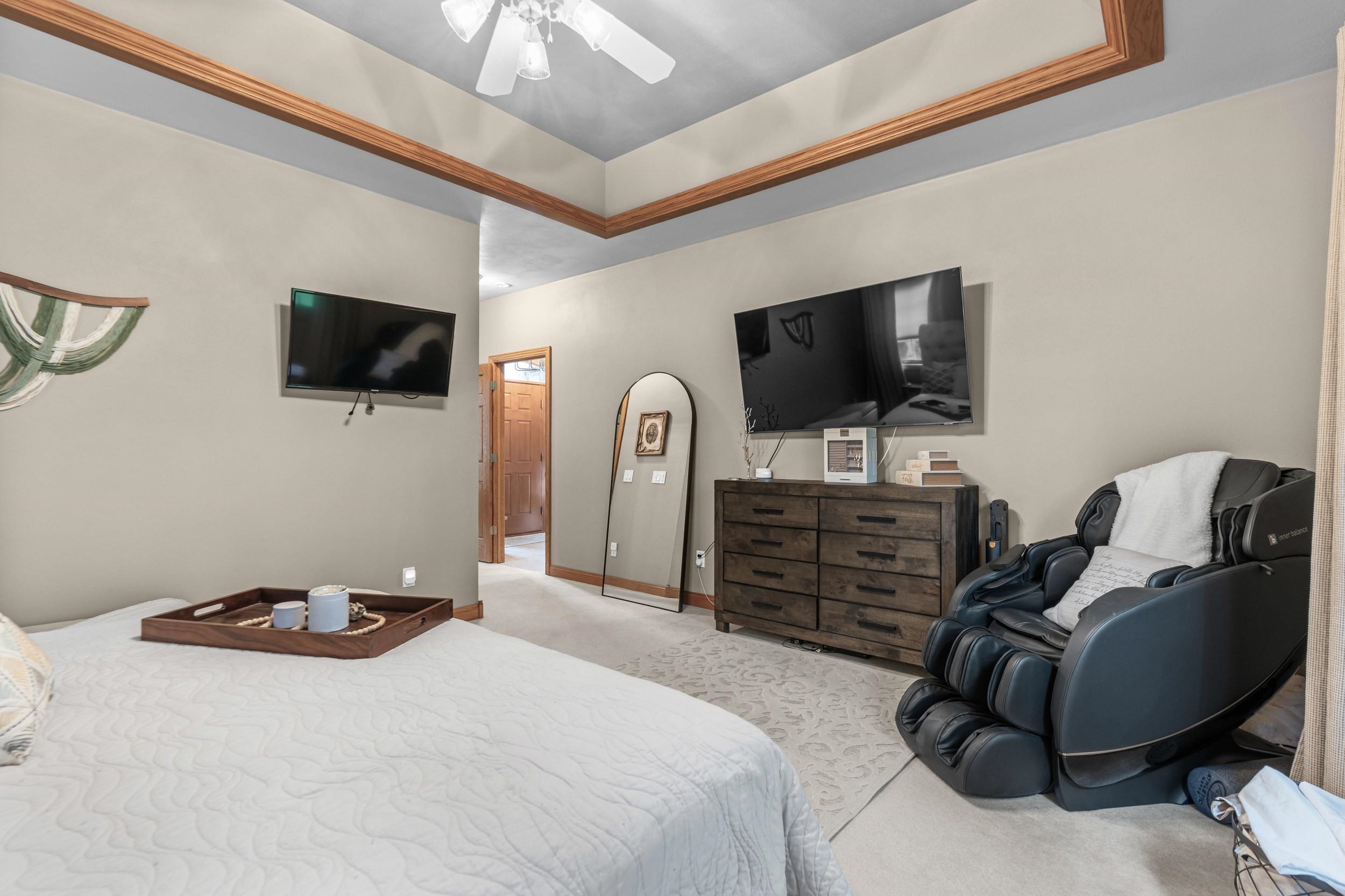 Furnished bedroom with a tray ceiling, two televisions, and a black massage chair.