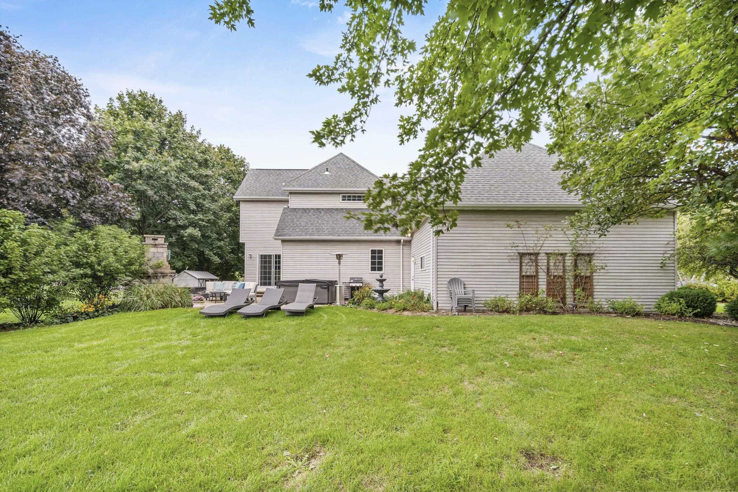 Rear of a two-story home beyond a broad lawn, with patio loungers and mature trees.