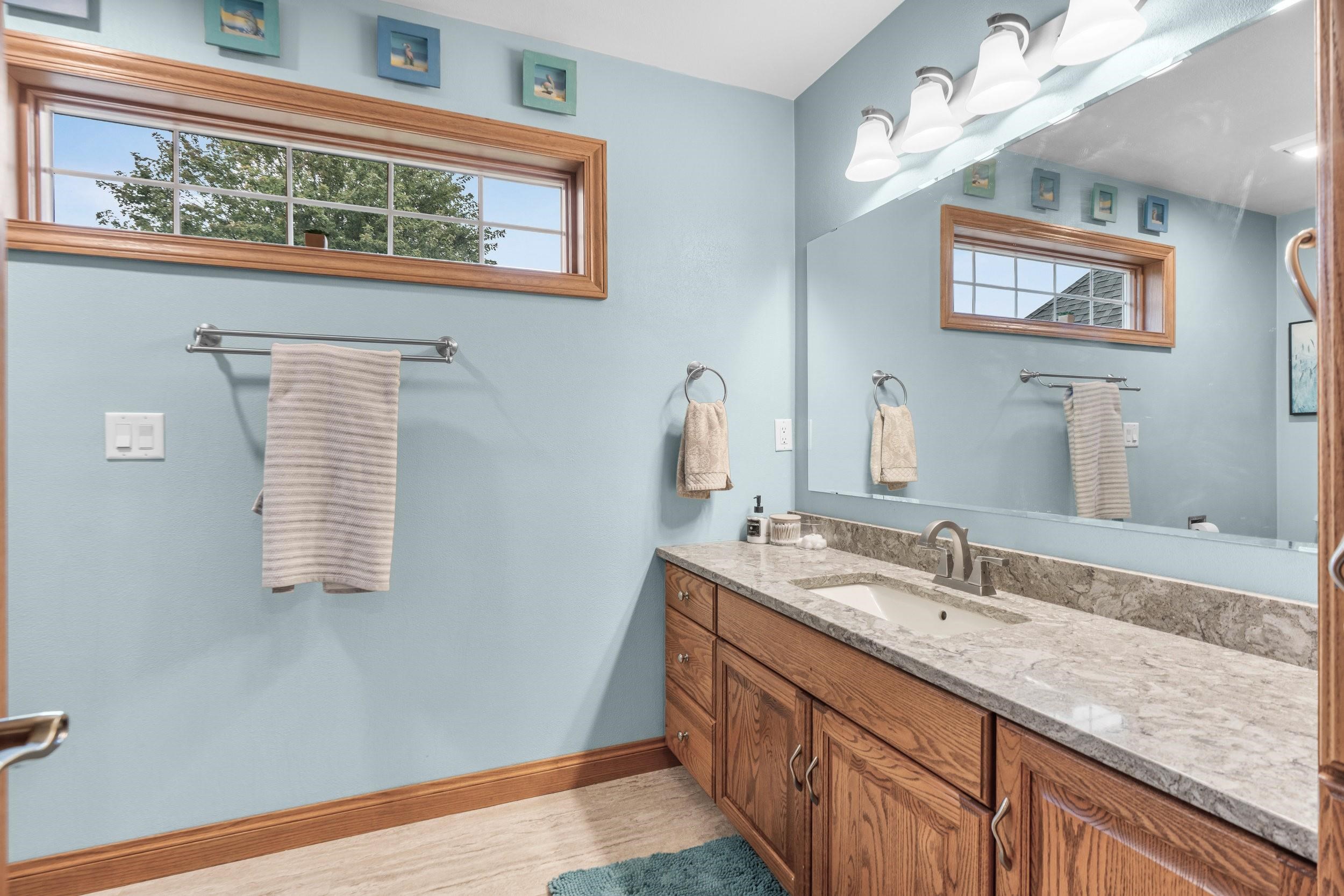 Bathroom vanity with a gray stone countertop, large mirror, and high wood-trimmed windows.