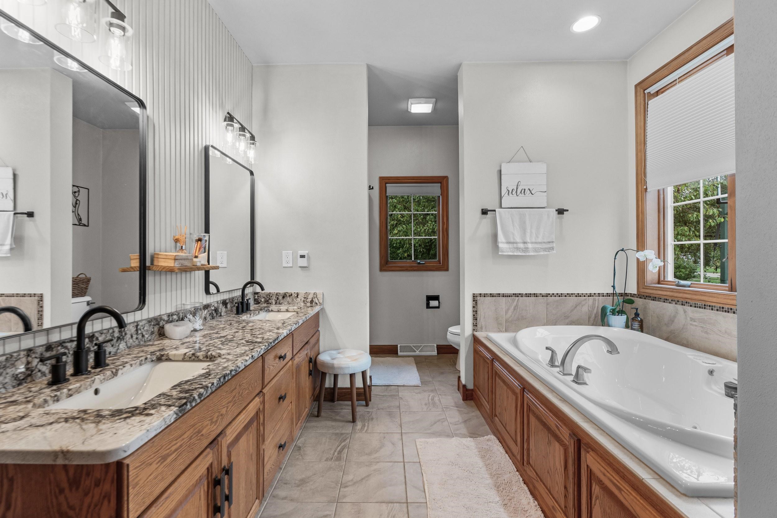 Bathroom with a long granite-topped vanity and a built-in soaking tub beside wood-trimmed windows.
